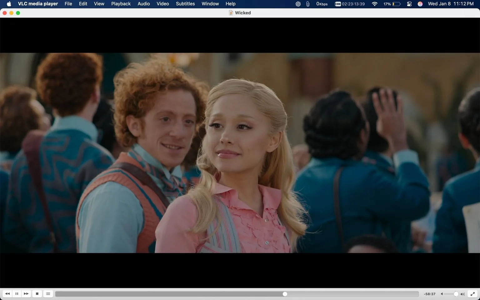 In the scene, a young woman with long, blonde hair styled in loose waves is prominently featured. She wears a pink blouse with ruffled details and a light blue vest, exuding a cheerful demeanor as she looks to her side. Behind her, a man with curly red hair and a playful expression appears to be engaging with her, dressed in a colorful outfit that includes a patterned vest and a bow tie. The background is filled with a crowd of people, many dressed in vibrant clothing, suggesting a lively outdoor event. The setting appears bright and festive, indicative of a celebratory atmosphere.