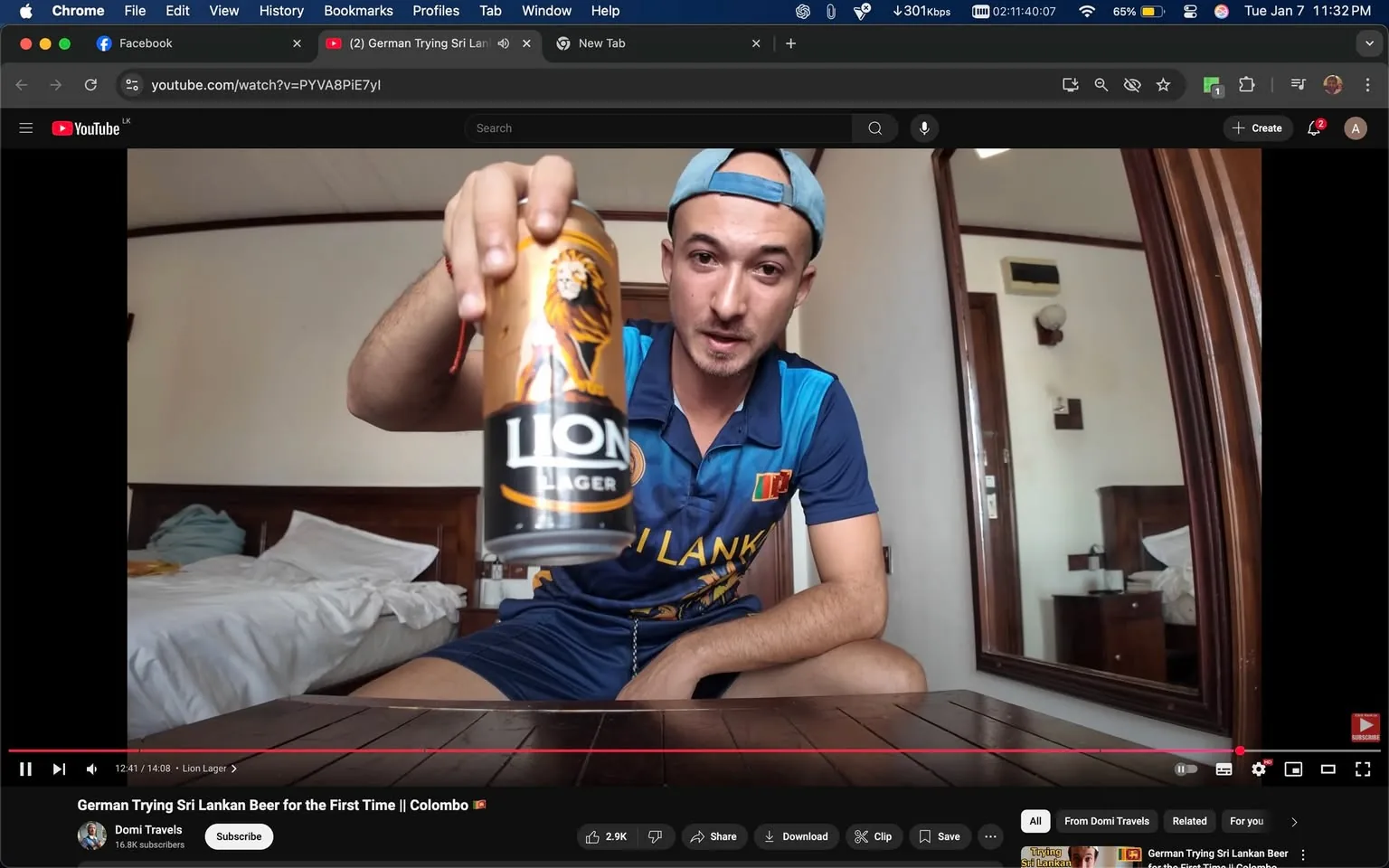 An unidentified adult is seated on the floor in a room, holding up a can of Lion Lager beer. The individual is wearing a blue sports jersey with Sri Lankan colors and a light blue cap. The background shows a bed with white sheets and a wooden headboard, along with a mirror reflecting part of the room. The setting appears casual and indoor, likely in a hotel or guesthouse. The can prominently displays the Lion Lager logo, which features a lion graphic. The person seems to be engaged in a video recording, possibly sharing their experience of trying Sri Lankan beer for the first time.