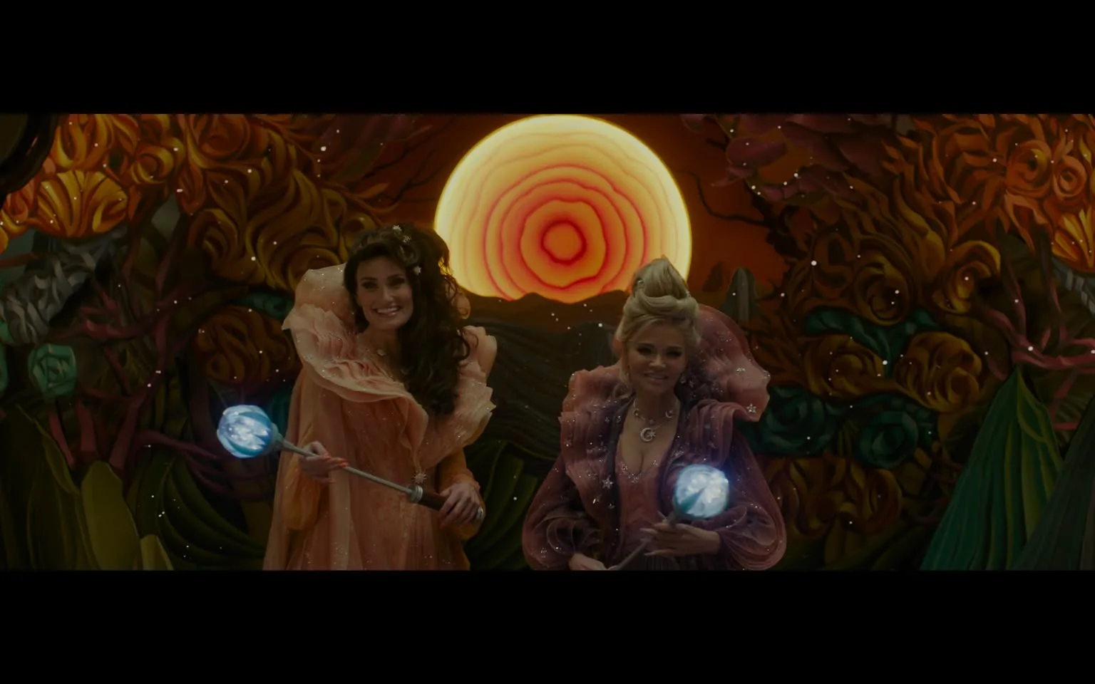 In the image, two women are prominently featured, both dressed in vibrant, flowing costumes that appear to be made of soft, sheer fabrics. The woman on the left has long, dark hair and is holding a wand with a glowing blue orb at the tip, while the woman on the right has light-colored hair styled in an updo and is also holding a similar wand. They are smiling and seem to be in a joyful or celebratory mood. The background is a striking, colorful mural depicting a large, stylized sun with concentric rings, surrounded by swirling, organic shapes in warm colors. The setting appears to be whimsical and fantastical, likely part of a themed event or performance. There are no visible brands or logos in the image.