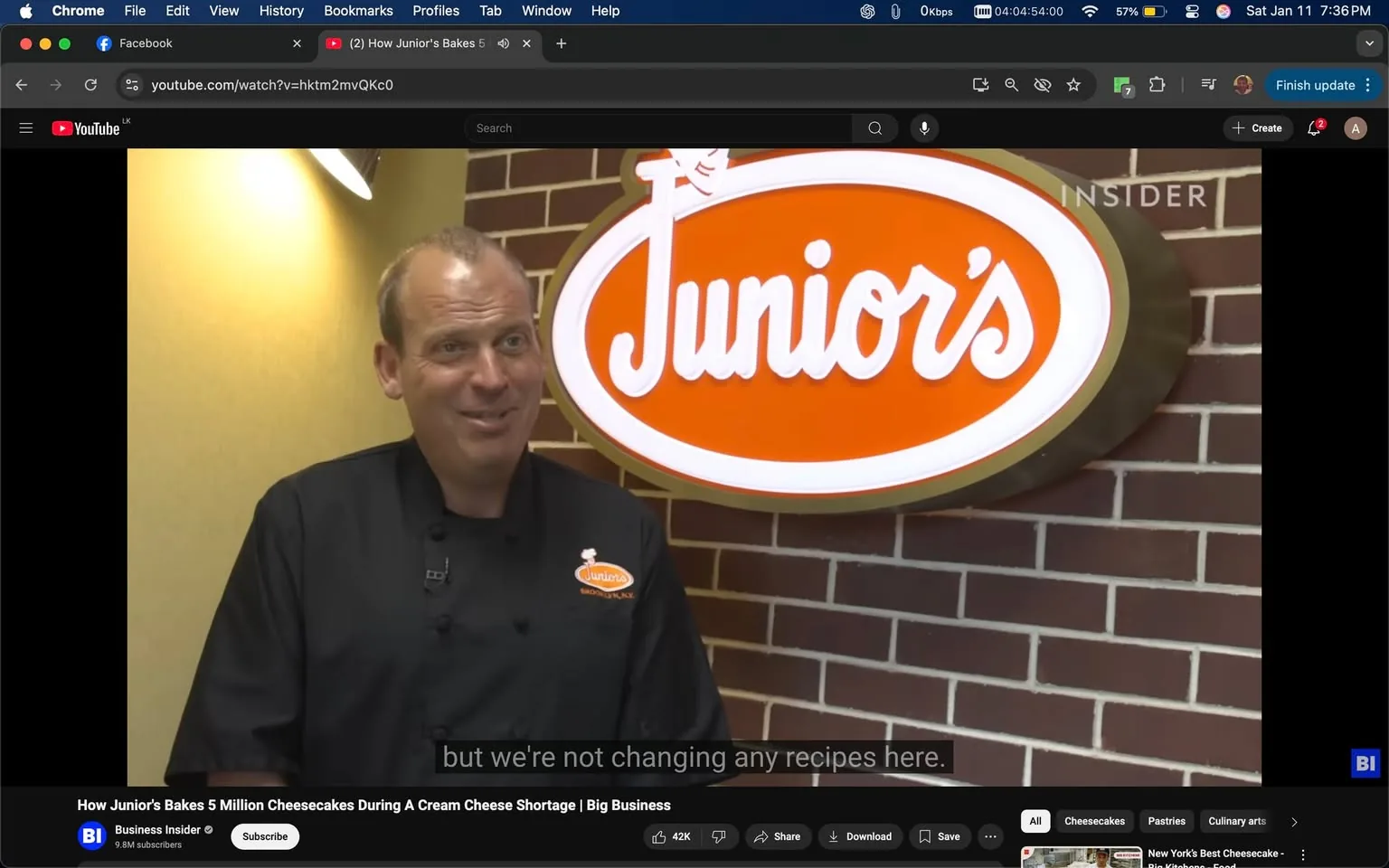 In the image, an unidentified adult male is speaking in front of a sign for "Junior's," a well-known brand associated with cheesecakes. He is wearing a black chef's jacket and appears to be in a casual indoor setting, likely a bakery or restaurant. The background features a brick wall, adding to the ambiance. The sign prominently displays the name "Junior's" in white lettering against an orange background. The individual has a friendly expression, suggesting a conversational tone as he discusses the brand's commitment to maintaining its recipes despite challenges, such as a cream cheese shortage. The video context indicates a focus on the business operations of Junior's.
