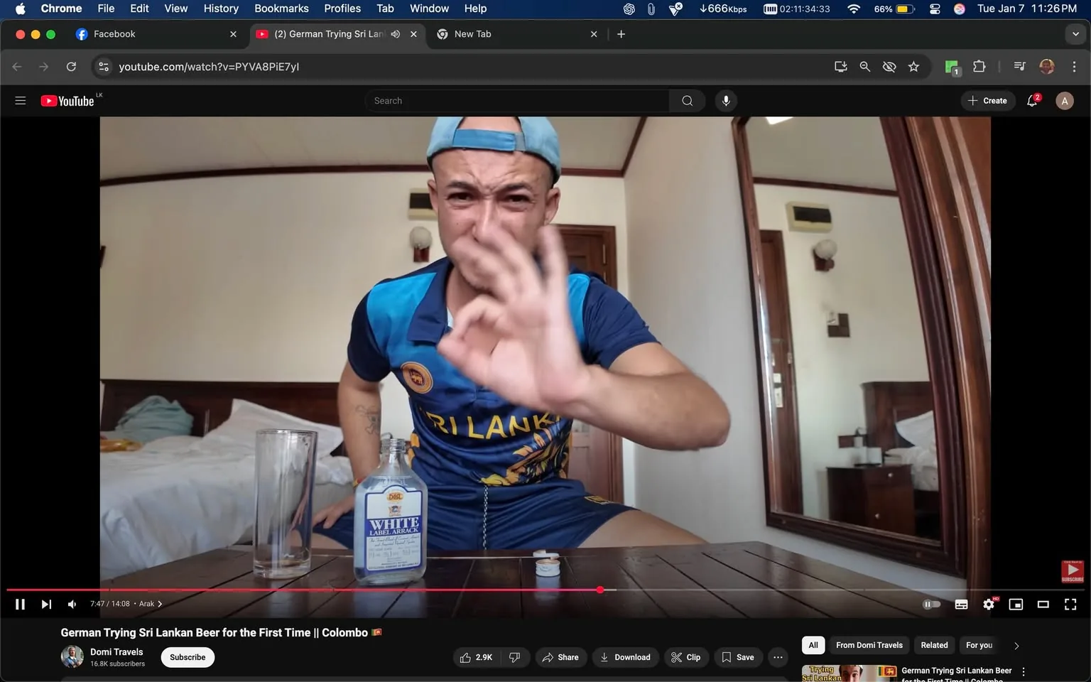 In the image, an unidentified adult male is seated at a wooden table in a room that appears to be a hotel or guesthouse. He is wearing a blue sports jersey with "SRI LANKA" printed on it and a light blue cap. The individual is making a hand gesture, possibly indicating a positive reaction, while looking directly at the camera with an expressive face. On the table in front of him is a clear glass and a bottle labeled "White Label Arak," suggesting he is preparing to try this alcoholic beverage. The background features a bed with white linens and wooden furniture, indicating an indoor setting. The overall atmosphere seems casual and relaxed, likely during a video recording for a social media platform.