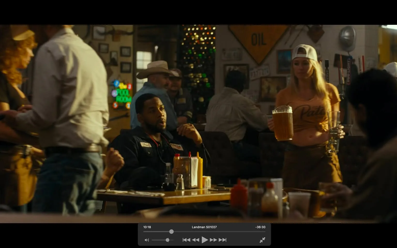In a lively bar setting, several individuals are present, engaged in conversation and activities. An unidentified adult male sits at a table, wearing a dark outfit with patches, appearing relaxed as he interacts with others. Nearby, a female server, dressed in a casual orange shirt and shorts, carries a large pitcher of beer and a glass, suggesting a busy service atmosphere. The background features various patrons, some wearing hats, and a colorful neon sign, adding to the vibrant ambiance. The decor includes wall signs and framed pictures, typical of a casual dining establishment. The scene conveys a sense of camaraderie and social interaction, with alcohol prominently featured.