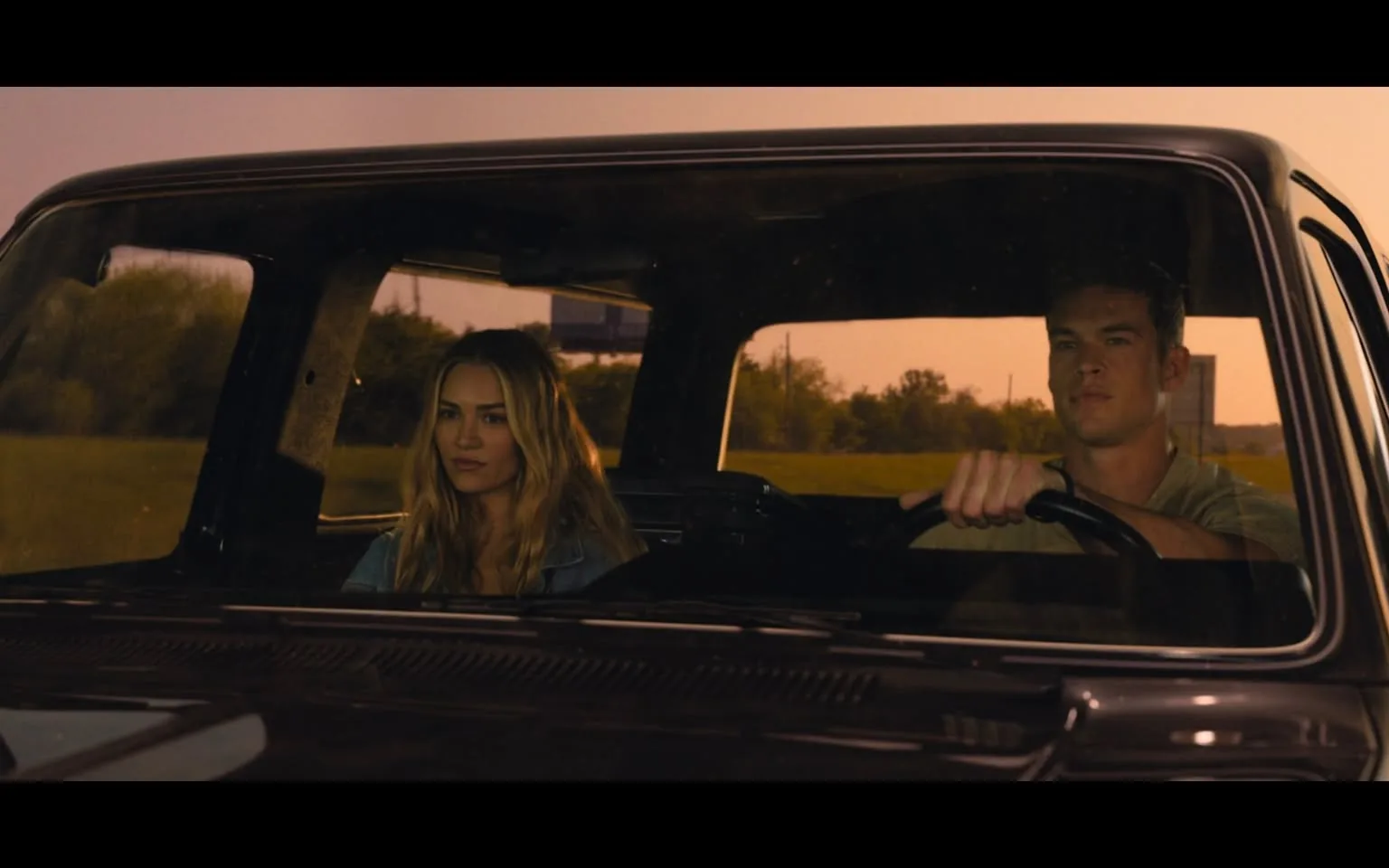 In the image, two individuals are seated inside a vehicle, with the driver on the right and a passenger on the left. The driver, a young adult male, is focused on the road, gripping the steering wheel with both hands. He has short hair and is wearing a light-colored shirt. The passenger, a young adult female, has long, wavy hair and is dressed in a denim jacket. The setting appears to be outdoors, with a soft, warm light suggesting either early morning or late afternoon. In the background, there are hints of greenery and a billboard visible through the passenger window, indicating a rural or suburban environment. The overall mood seems contemplative, as both individuals appear serious and engaged in their thoughts.