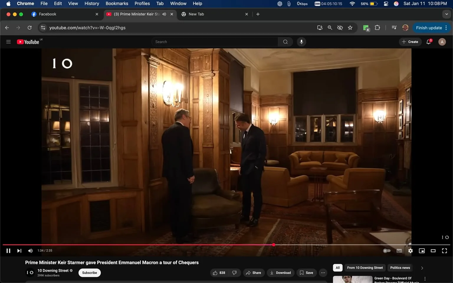 In a well-furnished room with wooden paneling and soft lighting, two individuals are engaged in conversation. One person, dressed in a dark suit, stands beside a green armchair, looking down at it. The other individual, also in formal attire, appears to be listening attentively. The setting features a large window with multiple panes, revealing a softly lit interior. In the background, there are several beige sofas arranged around a coffee table, and the walls are adorned with framed pictures. The atmosphere suggests a formal meeting or discussion, likely taking place in the evening given the warm lighting.