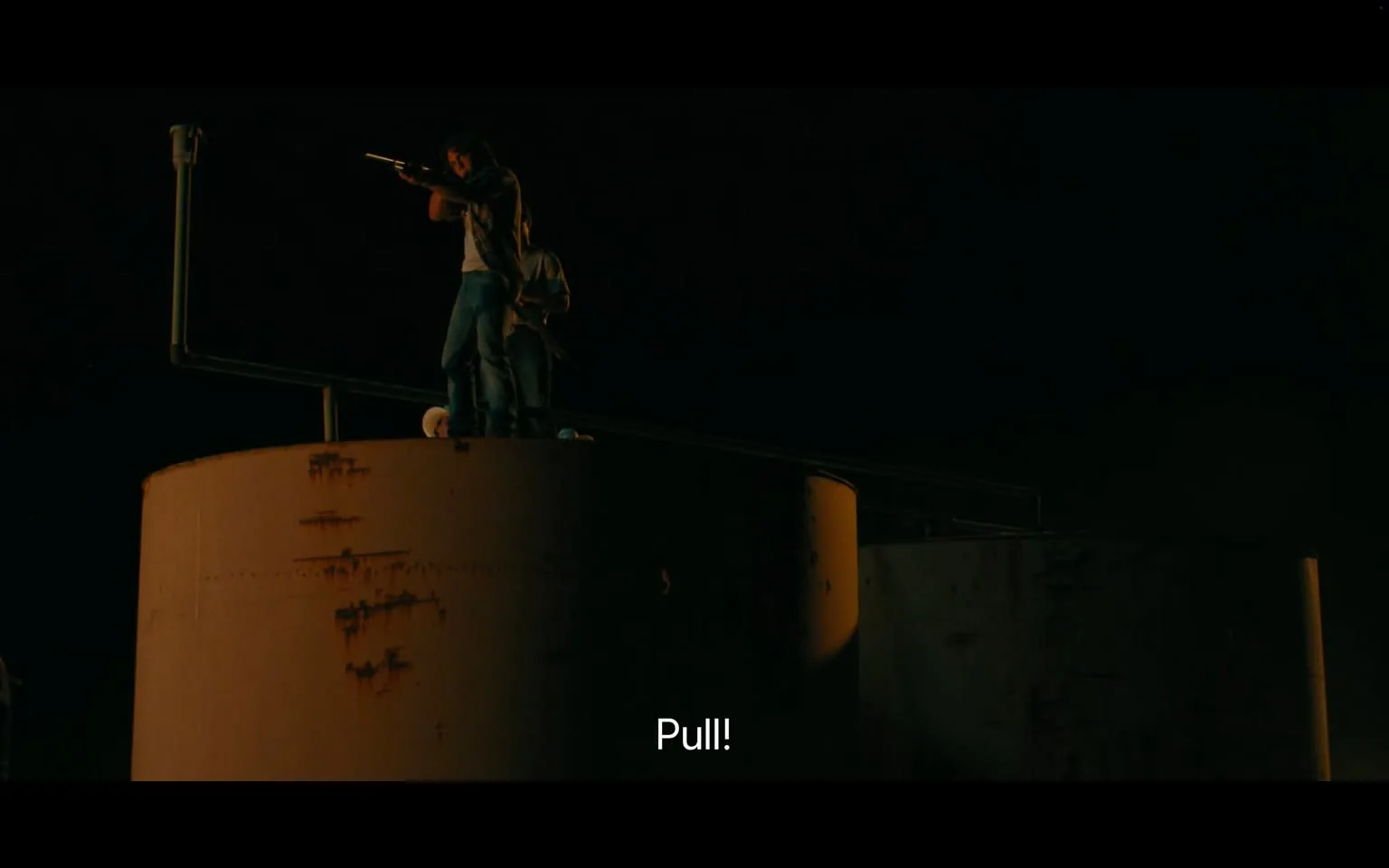 In a dimly lit outdoor setting at night, two individuals are positioned on top of a large, cylindrical structure, likely a tank. One person, standing with a rifle, appears focused and ready to take action, while the other is positioned behind them, possibly providing support. The structure shows signs of wear, with rust visible on its surface. The atmosphere suggests tension, as the person with the rifle is preparing to shoot, indicated by the command "Pull!" displayed prominently in the foreground. The scene conveys a sense of urgency and potential danger, with the dark surroundings enhancing the dramatic effect.