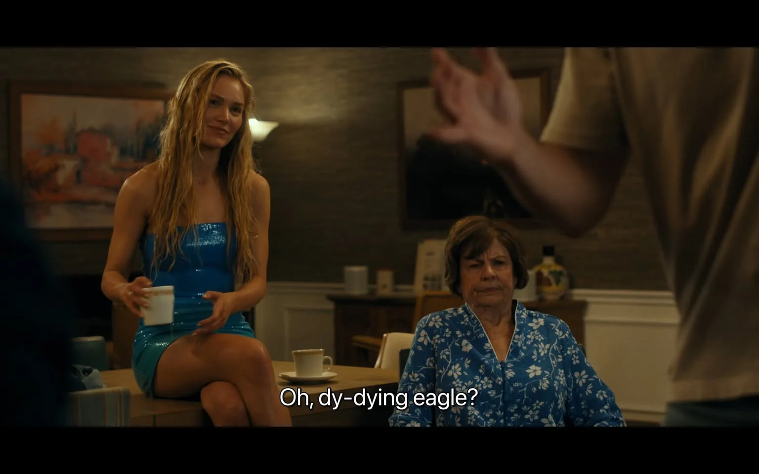 In the scene, a young woman with long, wavy hair is seated on a table, wearing a shiny blue dress and holding a white cup. She has a relaxed expression as she interacts with another woman, who is older and seated nearby. The older woman is dressed in a blue floral-patterned blouse and appears to be listening intently. The background features a softly lit room with a painting on the wall and various items on a table, suggesting a casual gathering. The dialogue includes a playful remark, indicating a light-hearted conversation. The overall atmosphere seems informal and friendly.