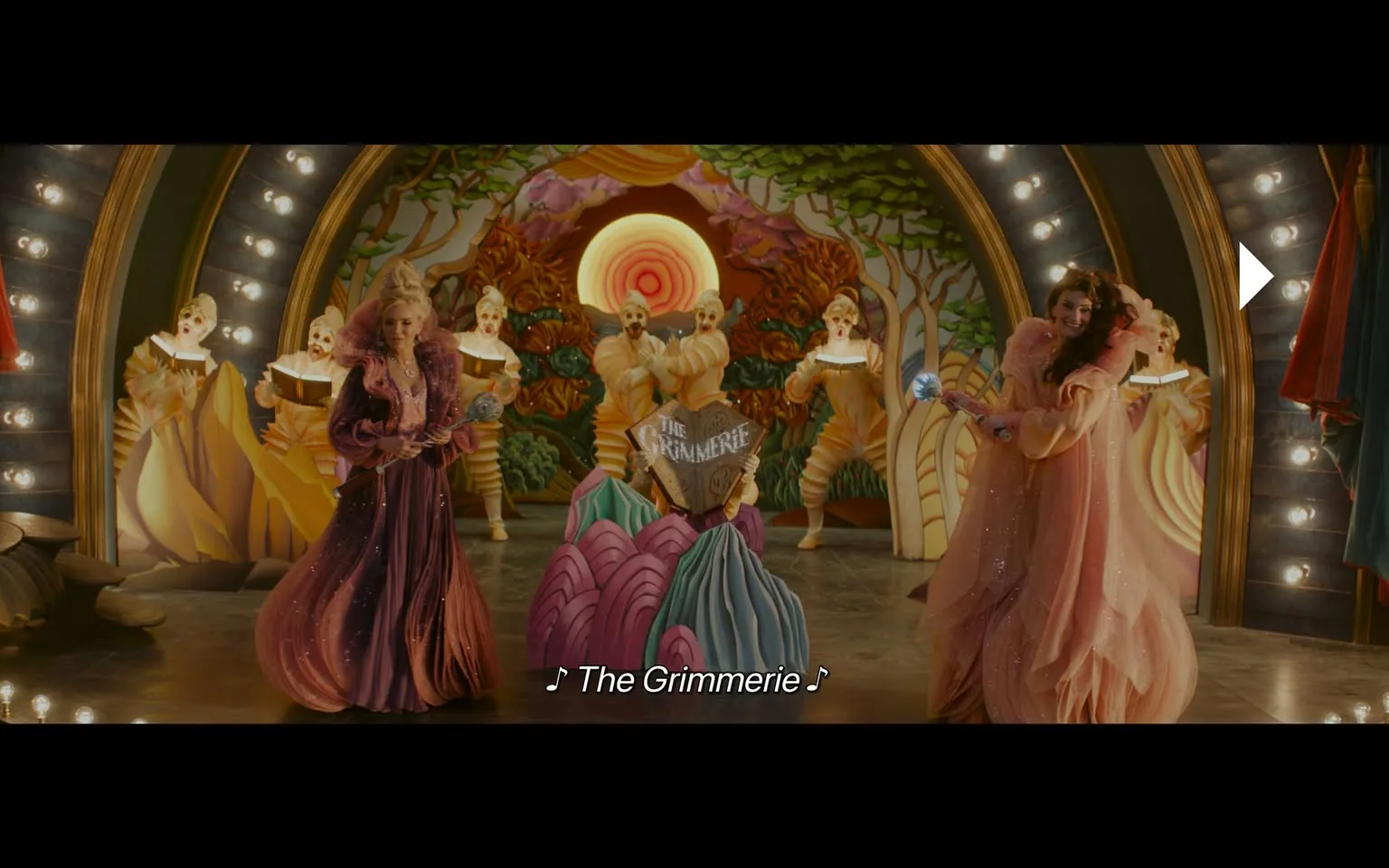 In a vibrant theatrical setting, two performers are prominently featured in elaborate costumes. One is dressed in a flowing, sparkly pink gown, while the other wears a rich purple ensemble adorned with jewels. They are positioned in front of a large, decorative backdrop that includes stylized trees and a sun motif, creating a whimsical atmosphere. Behind them, several figures in striped costumes appear to be part of a chorus, holding books and singing. The scene is lively, with the performers engaging with each other, and the text "The Grimmerie" is visible on a central prop, suggesting a musical or theatrical theme. The lighting is warm and inviting, enhancing the fantastical ambiance of the performance.