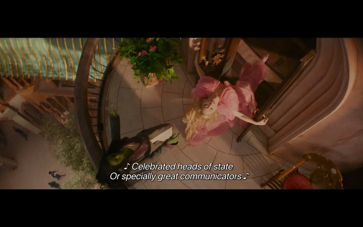 In the scene, two characters are depicted in a vibrant, whimsical environment. One character, dressed in a flowing pink outfit, appears to be suspended or gracefully leaning back, with long blonde hair cascading around her. The other character, dressed in dark clothing with green skin, is positioned below her, looking up. The setting features a spiral staircase and a balcony adorned with plants and flowers, suggesting an enchanting atmosphere. The background includes hints of a bustling outdoor scene, with people visible in the distance. The characters are engaged in a musical moment, as indicated by the lyrics displayed at the bottom of the image. The overall tone is playful and fantastical, characteristic of a musical or animated film.