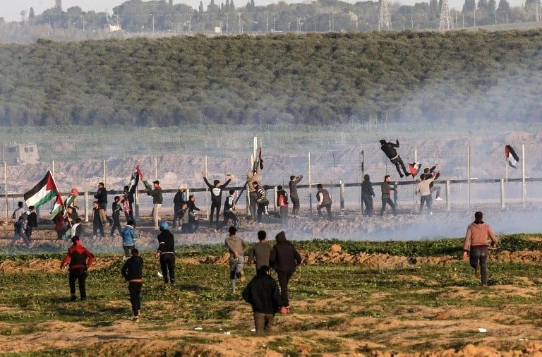 In a tense outdoor setting, a group of individuals is engaged in a protest, with many waving flags that feature a distinct color pattern, likely representing a political cause. The scene is chaotic, with some individuals appearing to throw objects, while others are running or standing amidst a cloud of smoke, suggesting the use of tear gas or similar crowd control measures. The protesters are dressed in a mix of casual clothing, with some wearing jackets and others in lighter attire, indicating a range of ages among the participants. The background features a barbed wire fence, which adds to the sense of conflict. The landscape is rural, with fields visible, and the atmosphere is charged with tension, reflecting the ongoing unrest.