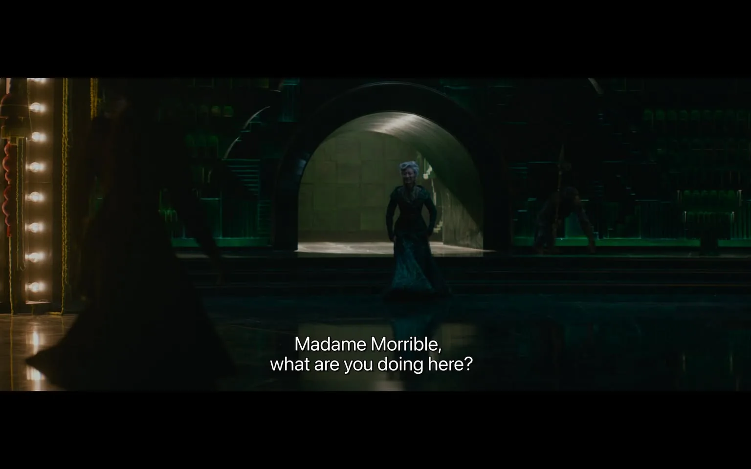 In the scene, a character dressed in a dark, elegant outfit stands in a dimly lit, dramatic setting, characterized by green lighting and architectural elements that suggest a theatrical environment. The character appears to be addressing another figure, referred to as "Madame Morrible," with a tone of inquiry. The background features an archway leading to a larger space, and there are hints of stairs and seating, indicating a venue designed for performances. The atmosphere is tense, likely reflecting a pivotal moment in the narrative. The dialogue suggests a confrontation or significant interaction between the characters.