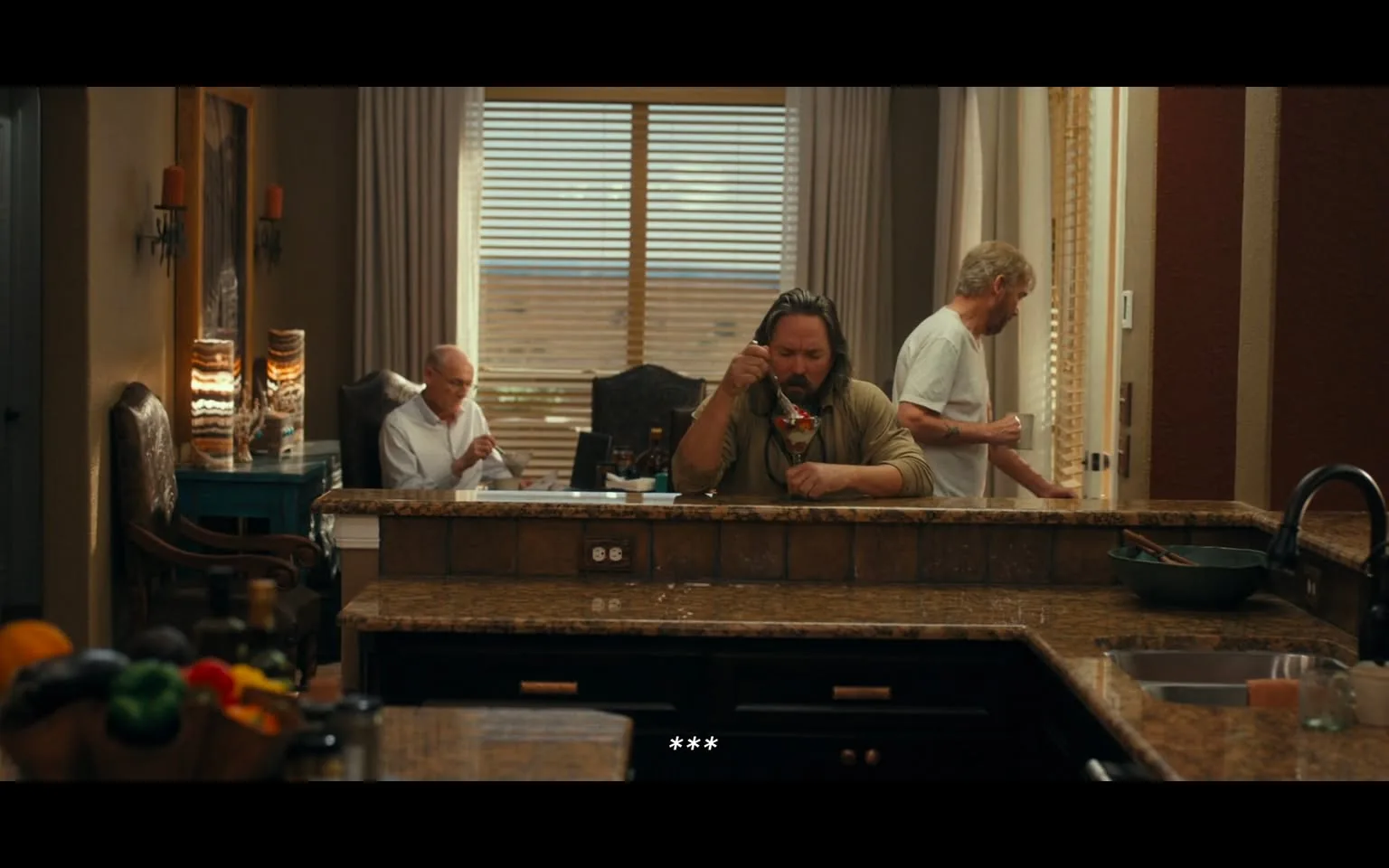 In a domestic kitchen setting, three unidentified adults are present. The central figure, seated at a granite countertop, is eating from a bowl, appearing to enjoy a meal. He has long hair and is dressed in a casual, long-sleeved shirt. To his left, another adult is seated at a table, focused on a document or meal, while a third adult stands in the background, holding a cup and looking towards the window. The kitchen features warm tones, with wooden cabinets and a variety of fruits visible in a bowl on the counter. Natural light filters through window blinds, illuminating the space. The atmosphere seems relaxed, with no visible signs of conflict or distress.