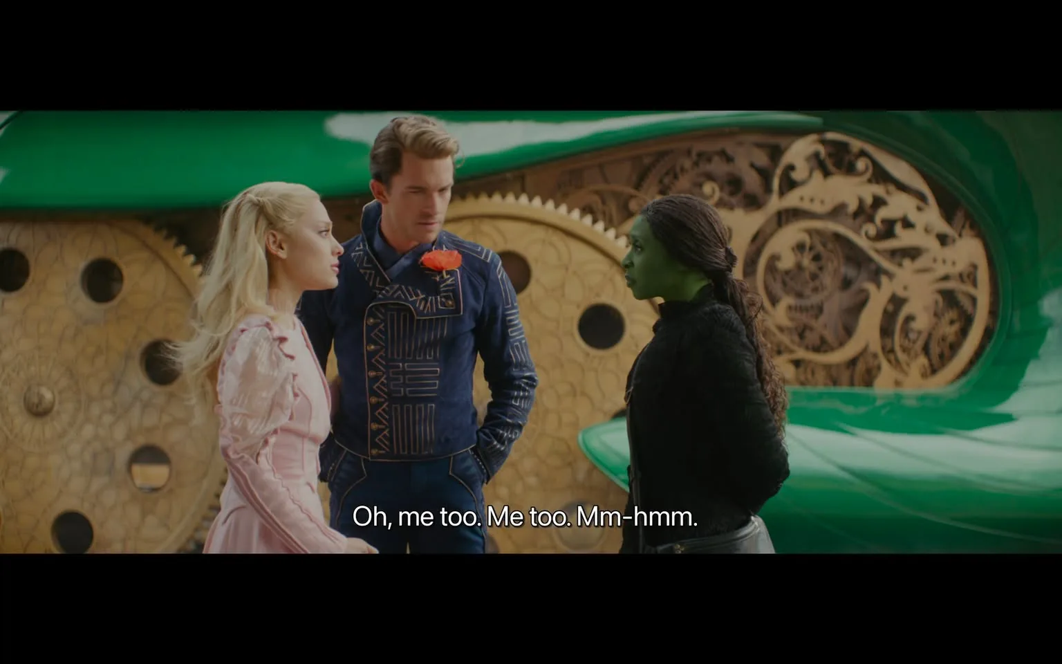 In the scene, three characters are engaged in conversation. One character, a woman with long blonde hair, is wearing a light pink dress with puffed sleeves. Next to her, a man in a blue outfit adorned with a geometric pattern and a red flower on his chest appears to be speaking. The third character, a woman with green skin and dark hair, is dressed in a black sweater and appears to be listening intently. The background features a large, green object with intricate designs, suggesting a fantastical or sci-fi setting. The dialogue displayed at the bottom indicates a casual exchange among the characters.