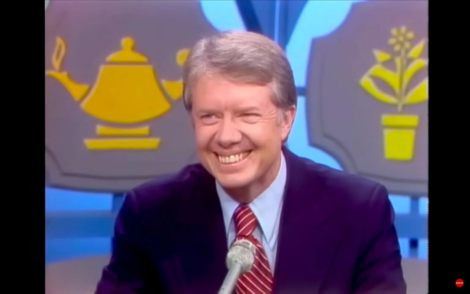 In the image, a smiling individual is seated at a table, dressed in a dark suit with a light blue shirt and a red striped tie. The background features two large, colorful icons: one resembling a teapot and the other depicting a potted plant, both in yellow against a gray backdrop. The setting appears to be a television studio, likely during a talk show or interview, given the formal attire and the presence of a microphone on the table. The individual exudes a friendly demeanor, suggesting a light-hearted or engaging conversation.