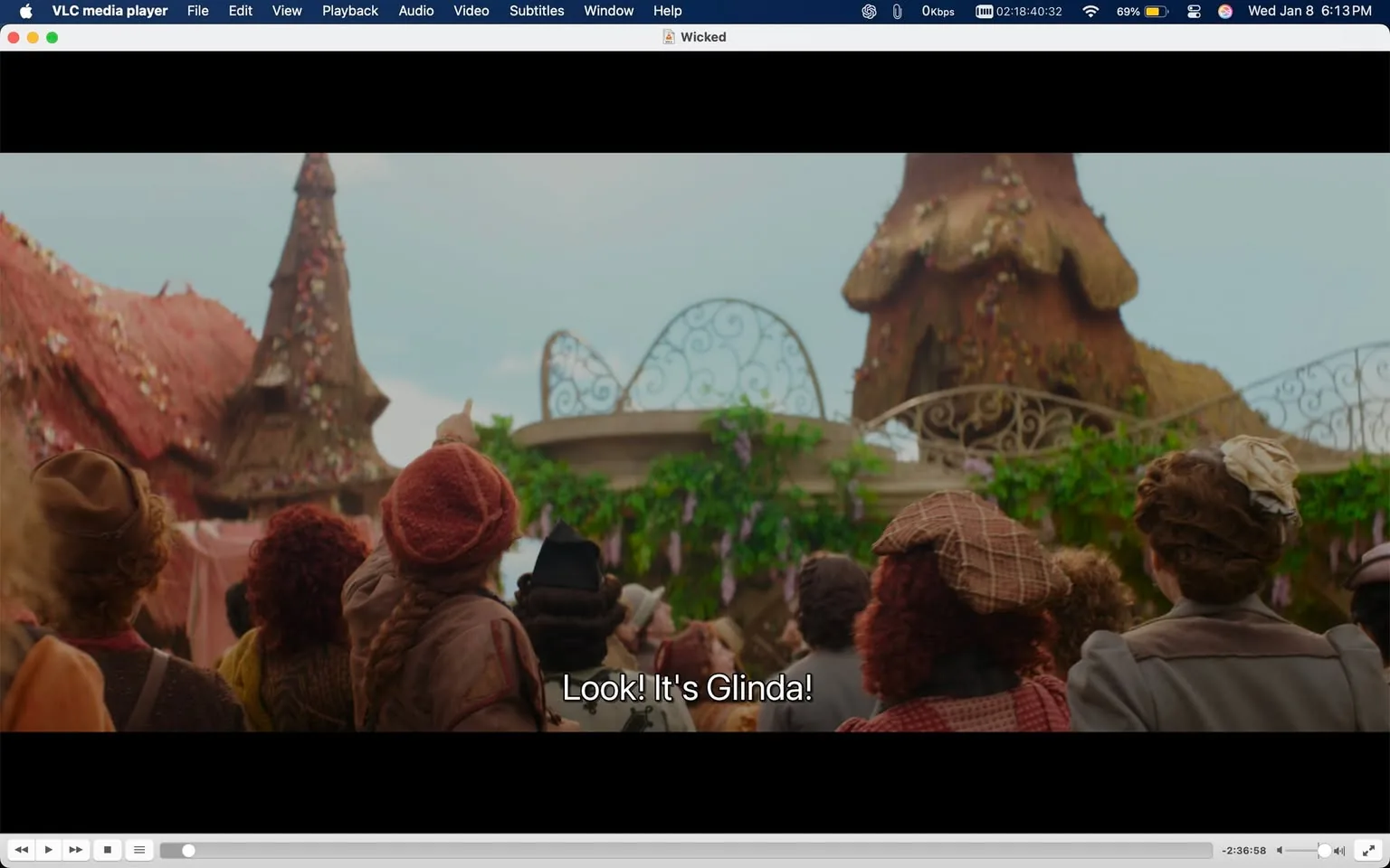 A crowd is gathered outdoors, looking towards a central figure, presumably Glinda, as indicated by the text overlay that reads, "Look! It's Glinda!" The people in the foreground are dressed in a variety of colorful, whimsical clothing, suggesting a festive atmosphere. Some individuals have curly hair, while others wear hats or headpieces. The background features whimsical architecture with a thatched roof and decorative elements, including flowers and greenery, contributing to a vibrant setting. The sky appears bright, indicating daytime. The expressions of the crowd suggest excitement or anticipation as they point and gaze towards the figure of Glinda.