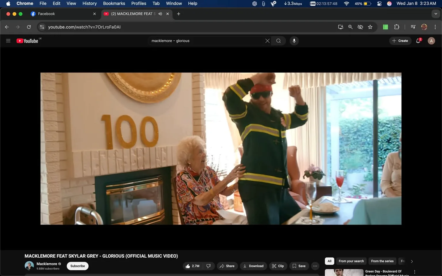 In a vibrant indoor setting, an unidentified adult is dancing joyfully, wearing a firefighter-style jacket with reflective stripes and a red bandana. They appear to be celebrating, possibly at a party, as indicated by the festive atmosphere. Nearby, an elderly woman, with curly white hair, is seated and smiling, seemingly enjoying the performance. The background features a fireplace and a decorative wall with the number "100" prominently displayed, suggesting a milestone celebration. The table is set with drinks and food, enhancing the celebratory mood. The overall scene conveys a sense of joy and togetherness among the participants.