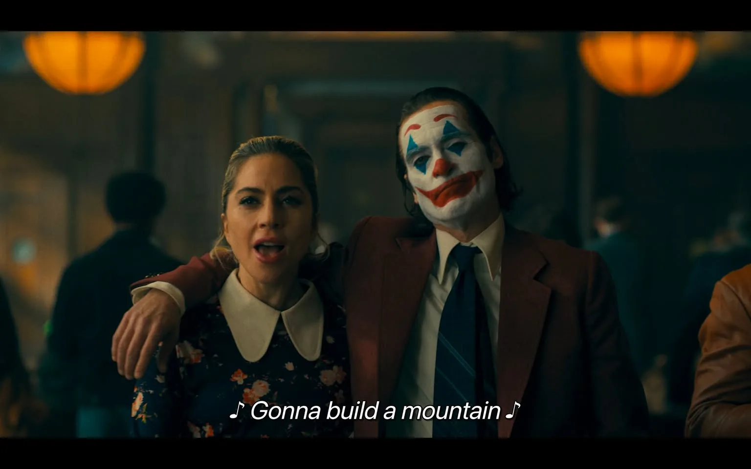 In the scene, two individuals are singing together, appearing to enjoy a lively moment. One person, dressed in a floral-patterned dress with a white collar, has a joyful expression as she sings. The other individual, wearing a red suit and clown makeup, has a painted face with blue and red accents, contributing to a whimsical atmosphere. The background suggests an indoor setting, possibly a gathering or event, with warm lighting from hanging lamps. The lyrics "Gonna build a mountain" are visible, indicating they are performing a song. The overall mood appears celebratory and engaging, with other people present in the background, though their faces are not clearly visible.