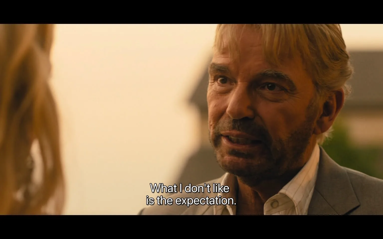 In the scene, an unidentified adult male is engaged in a serious conversation, expressing his feelings about expectations. He has a well-groomed appearance, with light-colored hair and a beard, wearing a light-colored suit over a white shirt. His facial expression is intense, suggesting he is passionate about the topic at hand. The background is softly blurred, indicating an outdoor setting during what appears to be sunset, with warm tones in the sky. The text overlay reads, "What I don't like is the expectation," emphasizing the man's emotional stance.