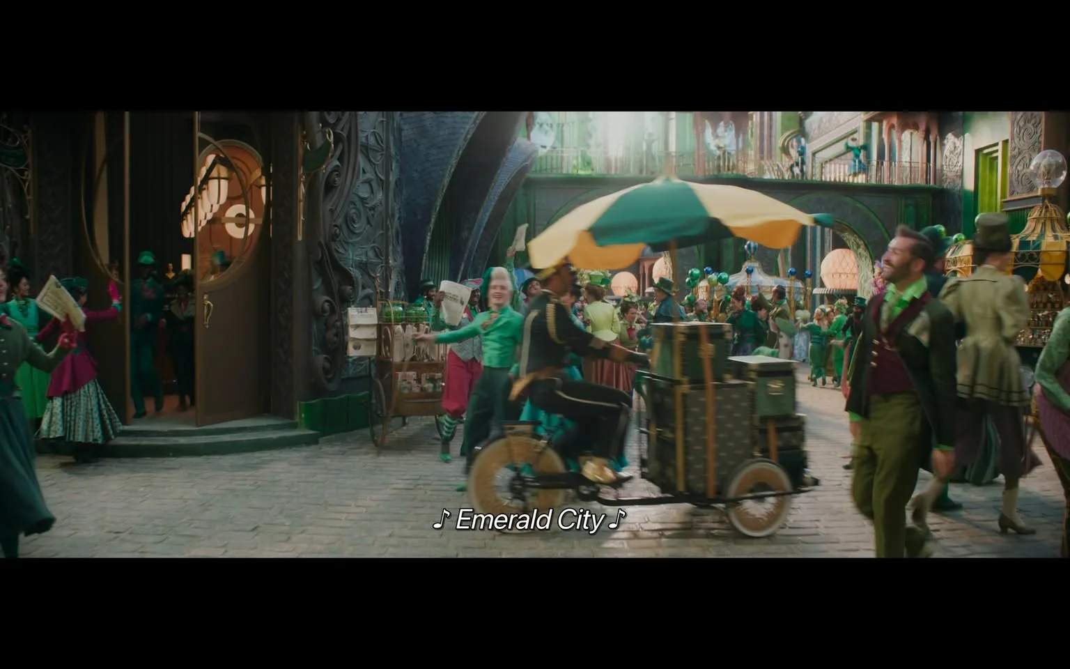 In a lively and colorful setting reminiscent of a fantastical city, a bustling street scene unfolds. People dressed in various shades of green and vibrant attire are engaged in activities, with some holding newspapers and others interacting with a vendor. A man rides a bicycle-like cart adorned with a large green and yellow umbrella, suggesting a festive atmosphere. The architecture features ornate designs, with a prominent circular window displaying intricate patterns. The scene is filled with joy and movement, as individuals appear to be celebrating, possibly in a parade or festival. The background is rich with details, including decorative elements and additional characters, contributing to the overall whimsical ambiance.
