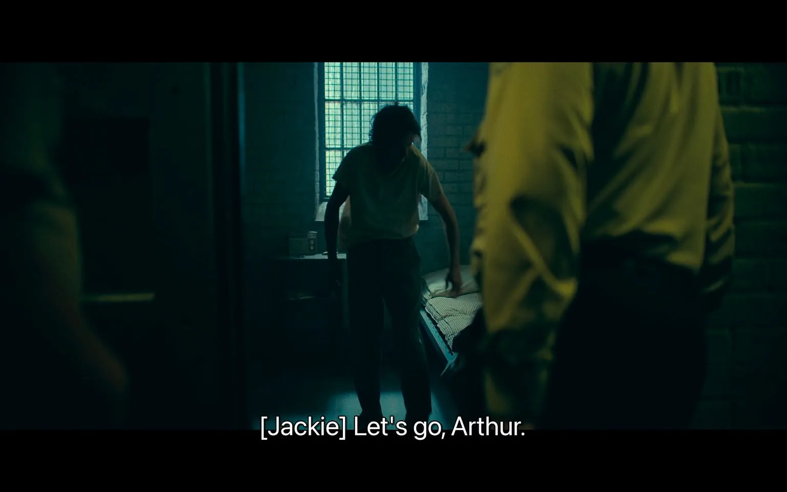 In a dimly lit room resembling a prison cell, an unidentified adult male is seen standing near a bed, dressed in a simple white shirt and dark pants. He appears to be in a state of distress or reluctance as he leans forward, possibly preparing to leave the space. The atmosphere is tense, accentuated by the presence of another individual, who is partially visible and wearing a yellow uniform, suggesting a guard or authority figure. The room features a barred window allowing limited light, casting shadows that enhance the somber mood. A small radio is visible on a shelf, adding to the sparse decor. The dialogue indicates a command to leave, hinting at a narrative of confinement and authority.