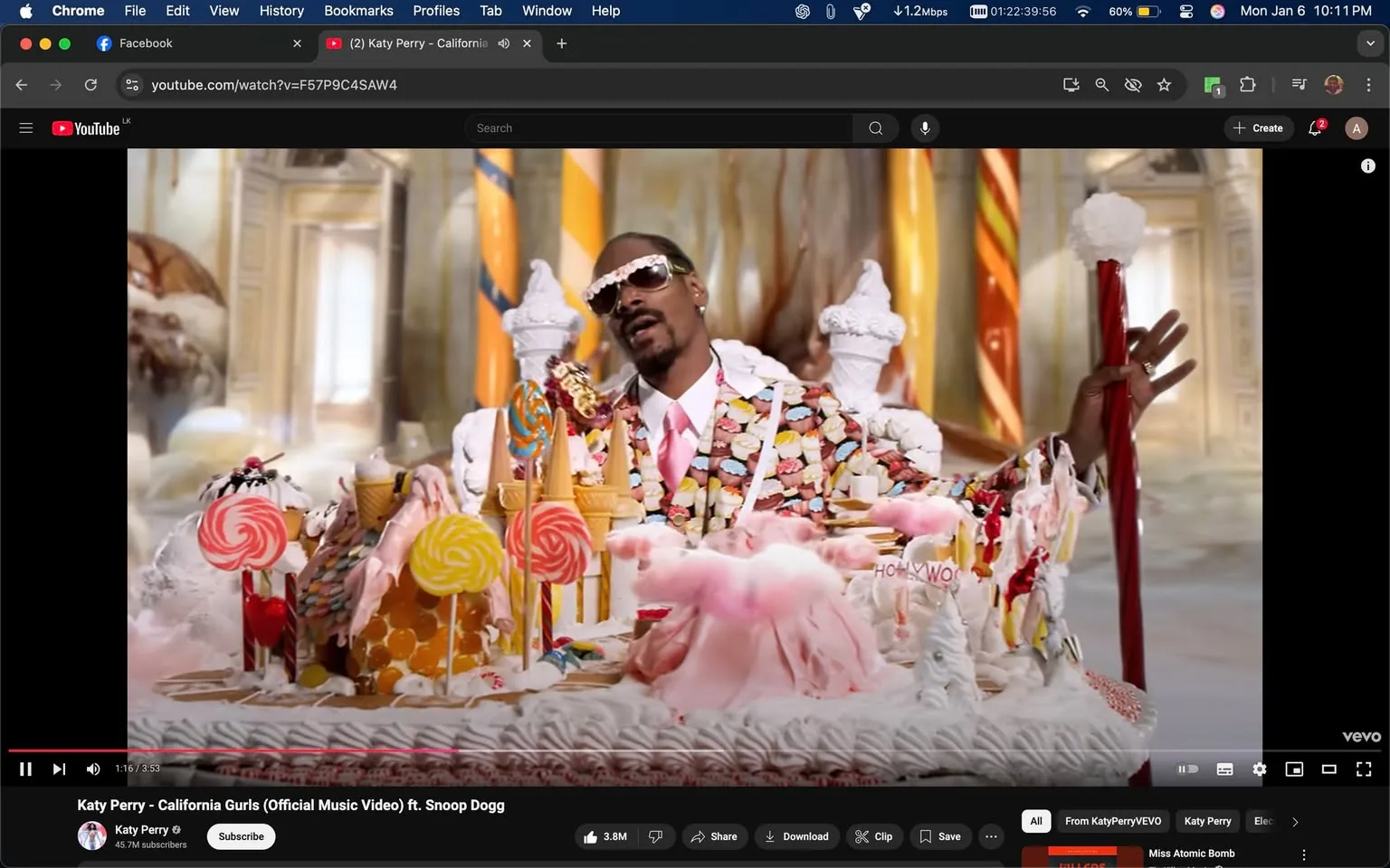 In a vibrant and whimsical setting, a person is seated amidst an elaborate display of candy-themed decorations. The individual is wearing a suit adorned with colorful candy patterns, complemented by sunglasses and a pink tie. Surrounding them are various sweets, including large lollipops, ice cream cones, and cotton candy, creating a playful and indulgent atmosphere. The background features ornate architecture with bright colors, enhancing the fantastical feel of the scene. The overall mood is lighthearted and celebratory, typical of a music video aesthetic.