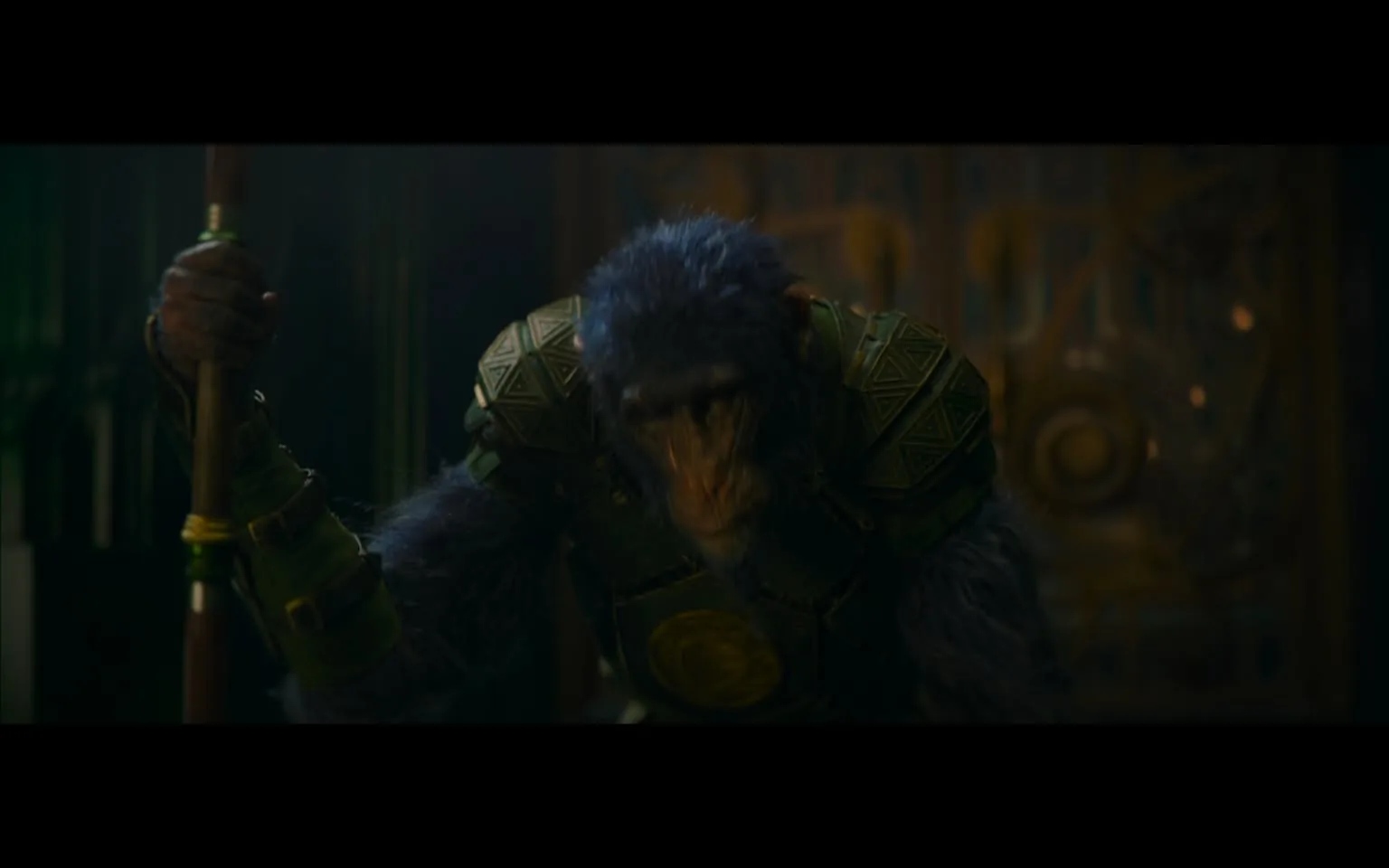 In the image, a humanoid ape character is depicted, wearing intricate armor adorned with geometric patterns. The armor appears to be made of a metallic material, with a prominent circular emblem on the chest. The character is holding a wooden staff, which adds a sense of authority or power. The setting is dimly lit, with hints of a fantastical environment in the background, featuring ornate designs that suggest an ancient or mythical theme. The character's expression is serious, and the overall atmosphere conveys a sense of tension or anticipation.