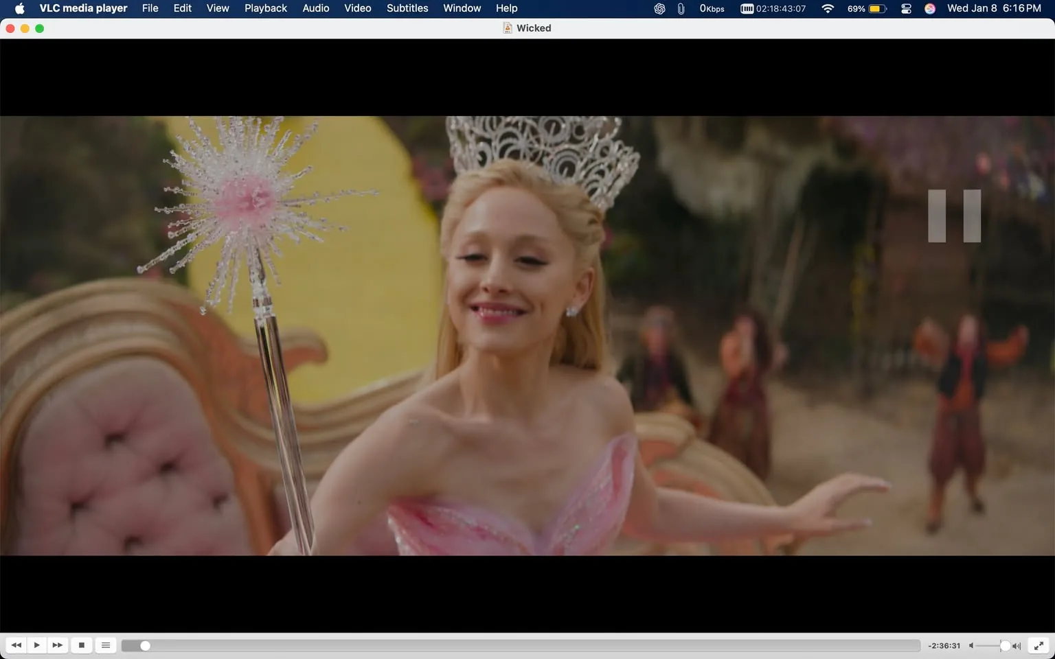 In a vibrant outdoor setting, a character dressed in a sparkling pink gown is joyfully interacting with her surroundings. She has long, blonde hair and is wearing a tiara, which adds to her regal appearance. In her hand, she holds a magical wand topped with a starburst design, suggesting a whimsical or fantasy theme. The background features blurred figures, possibly other characters, who appear to be engaged in a lively scene. The overall atmosphere is cheerful and enchanting, indicative of a performance or celebration. The setting is bright and colorful, enhancing the magical feel of the moment.