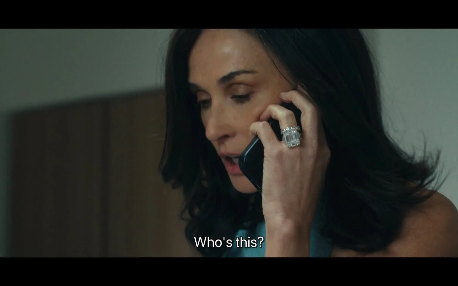 In the image, a woman is engaged in a phone conversation, appearing to be in a moment of concern or surprise as she asks, "Who's this?" She has long, dark hair and is wearing a light-colored top. Her expression suggests she is focused on the call, and she is holding a black phone to her ear with her right hand. On her left hand, she wears a large, ornate ring. The background is softly blurred, indicating an indoor setting, likely a home or office, with neutral-colored walls and furniture. The overall atmosphere conveys a sense of urgency or tension in the conversation.