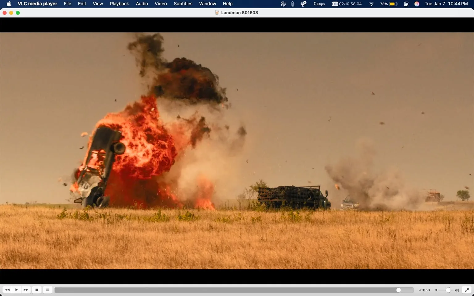 A vehicle is caught in a dramatic explosion, sending flames and debris into the air against a backdrop of a clear, dusty sky. The explosion is intense, with bright orange and red flames dominating the scene, while thick black smoke billows upward. In the foreground, a field of dry grass is visible, indicating an outdoor setting. Another vehicle can be seen in the background, possibly affected by the explosion. The overall atmosphere conveys a sense of chaos and destruction, typical of action sequences in films.