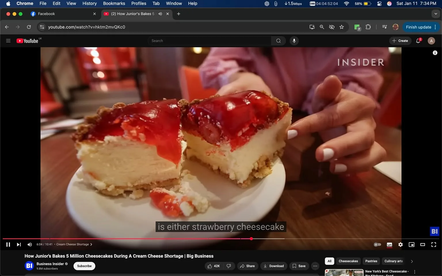 A slice of cheesecake is prominently displayed on a white plate, showcasing a creamy filling topped with a glossy layer of red strawberry sauce. The cheesecake appears to have a crumbly crust, likely made from graham crackers. A hand, possibly belonging to an unidentified adult, is reaching towards the dessert, with neatly manicured nails painted white. The background features a warm, indoor setting with soft lighting and hints of red decor, suggesting a cozy atmosphere. The visible text in the image mentions "strawberry cheesecake," indicating the flavor of the dessert.