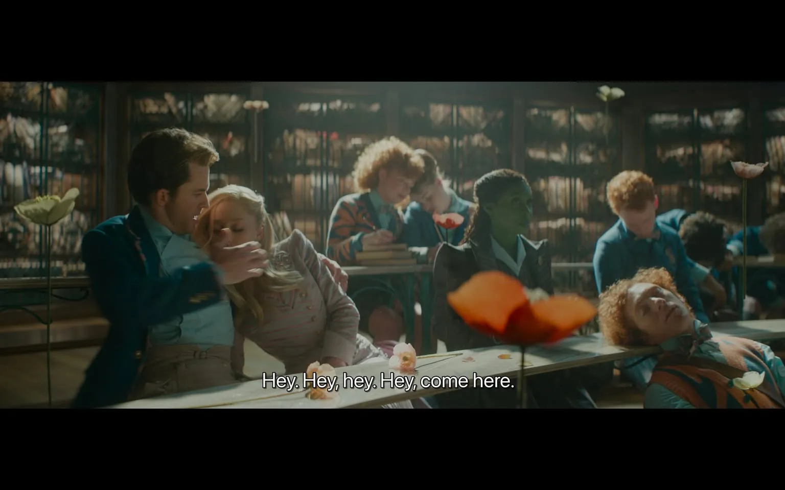 In a classroom filled with students, a young man leans in closely to a young woman seated beside him, appearing to whisper or speak to her. The woman, with long blonde hair, seems to be leaning towards him, suggesting a moment of intimacy or distraction. Other students in the background are engaged in their own activities; some appear to be resting their heads on their desks, while others are writing or looking at their books. The classroom has a warm, softly lit atmosphere, with decorative flowers visible on the desks, adding a whimsical touch to the setting. The students are dressed in a mix of formal and semi-casual attire, indicative of a school environment. The overall mood seems relaxed, with a hint of playful interaction among the students.