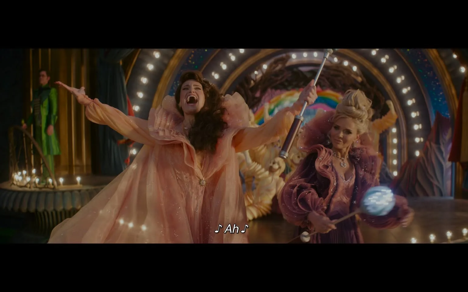 In a lively theatrical performance, two individuals are prominently featured. The first person, dressed in a flowing, sparkly pink gown with dramatic ruffles, is singing passionately, arms outstretched, conveying a sense of joy and exuberance. The second individual, wearing a rich purple outfit adorned with sequins, holds a staff topped with a glowing orb, suggesting a magical theme. The background is colorful and whimsical, with bright lights and a rainbow motif, enhancing the fantastical atmosphere of the scene. The setting appears to be a stage, likely part of a musical or performance event, with additional performers visible in the background.