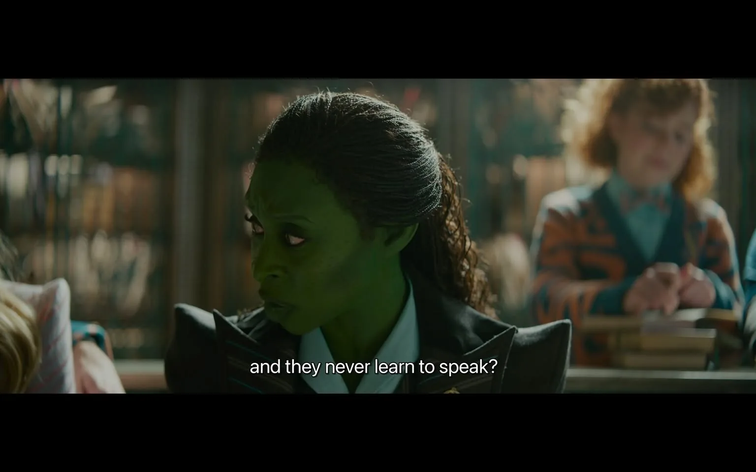 In the scene, a character with green skin and textured hair is engaged in a serious conversation, looking intently to the side. She wears a dark suit with a collar, suggesting a formal or professional setting. The background features blurred figures, including a person with curly hair and a colorful striped outfit, indicating a classroom or similar environment. The character's expression conveys curiosity or concern, as she poses a question about communication. The overall atmosphere appears to be one of discussion or inquiry, likely within a narrative context.
