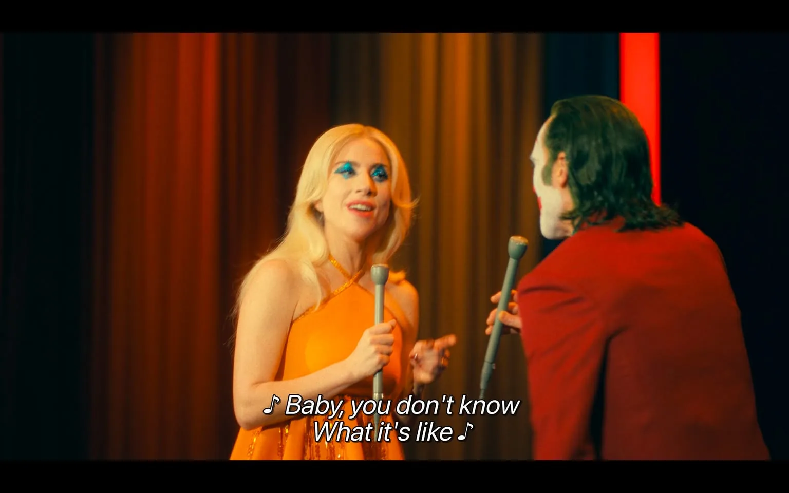 In a vibrant theatrical setting, two performers are engaged in a duet on stage. One performer, dressed in a bright orange outfit, has long, blonde hair and striking blue eye makeup, conveying a lively and expressive demeanor. The other performer, wearing a red suit and white face paint, faces the first performer, holding a microphone. The background features colorful curtains, enhancing the dramatic atmosphere of the performance. The lyrics displayed on the screen suggest an emotional theme, with the line "Baby, you don't know what it's like" indicating a connection between the performers. The overall scene captures a moment of artistic expression and interaction between the two individuals.