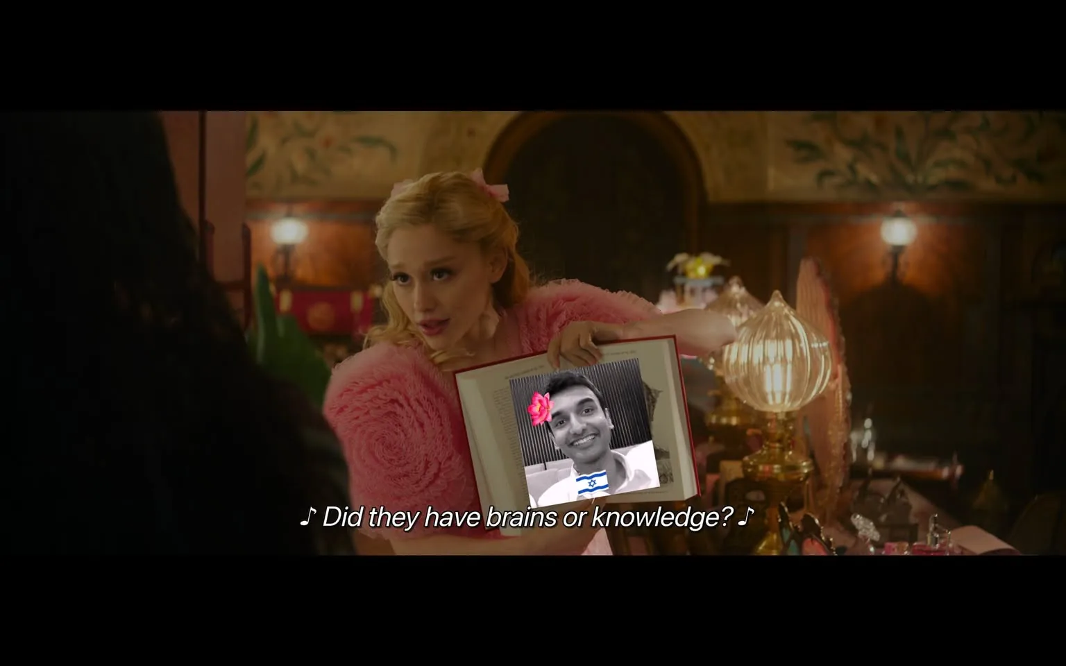 In the scene, a character with blonde hair styled in loose waves is wearing a fluffy pink sweater. She is holding a framed picture that appears to contain a photo of a man, who is smiling and has a flower graphic overlay. The background features ornate decor, including a lamp and various decorative items, suggesting an indoor setting with a vintage or whimsical aesthetic. The character is engaged in a performance, indicated by the musical notes and lyrics displayed at the bottom of the image. The overall atmosphere seems lively and theatrical.