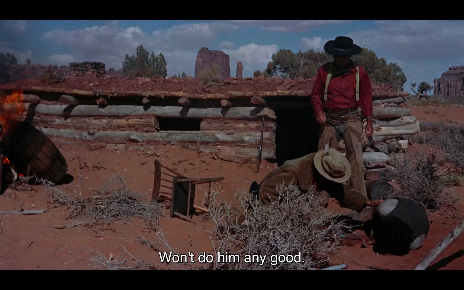 In a rugged outdoor setting typical of a Western film, two unidentified adults are present near a log cabin. One person, dressed in a red shirt and dark hat, stands over another who is crouched down, appearing to assist or confront them. The background features a barrel that is on fire, adding to the tension of the scene. A chair lies overturned on the ground, and a rifle is visible leaning against the cabin. The landscape includes sparse vegetation and rocky formations, suggesting a remote location. The dialogue, "Won't do him any good," hints at a serious or urgent situation. The overall atmosphere conveys a sense of conflict or danger.