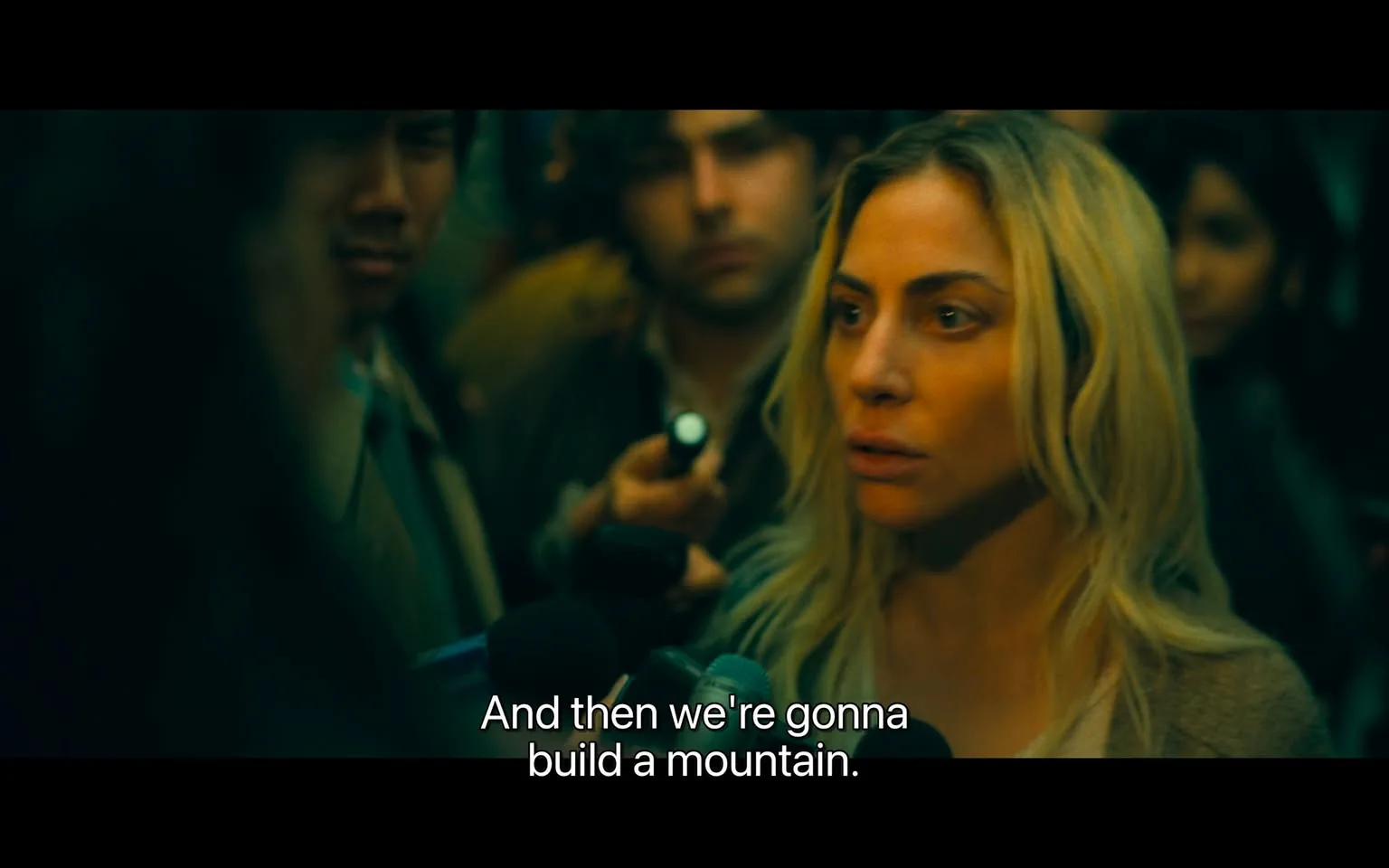 In a crowded setting, a woman with long, blonde hair is prominently featured, speaking intently into a microphone. Her expression conveys determination as she states, "And then we're gonna build a mountain." Surrounding her are several individuals, including a man with dark hair and a serious expression, and another person partially obscured by the crowd. The atmosphere appears charged, likely indicative of a press conference or public gathering. The lighting is dim, contributing to a sense of urgency or tension in the scene. No visible brands or logos are present, and there are no indications of violence or sexual content.
