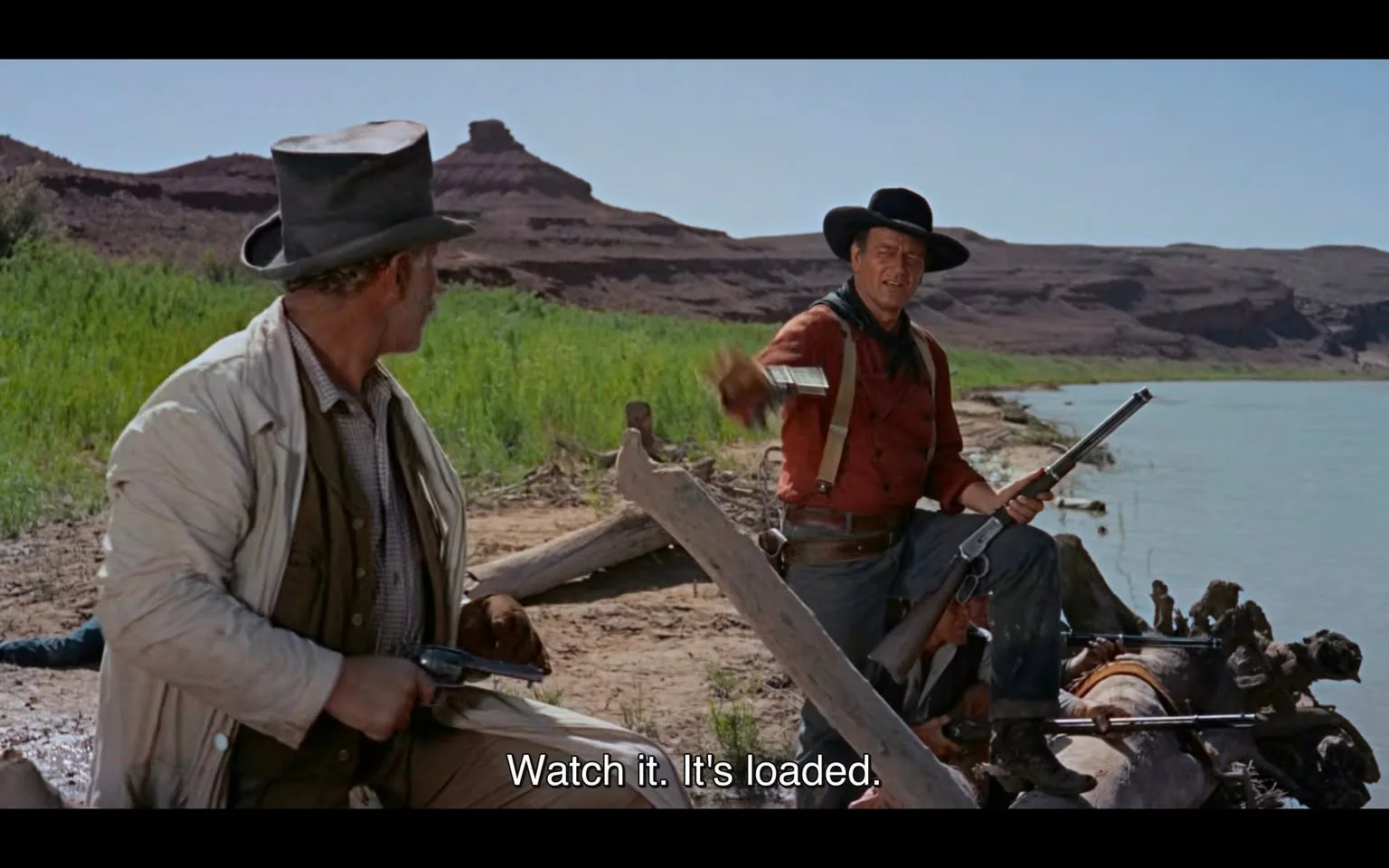 In the scene, two men are seated near a riverbank in a western landscape characterized by rugged terrain and sparse vegetation. One man, dressed in a red shirt and black hat, is holding a rifle and appears to be warning the other, who is wearing a light-colored coat and a dark hat, about the weapon being loaded. The second man has a revolver in his hand and looks towards the first, suggesting a tense interaction. The background features a calm river and distant hills, indicating a sunny day. The dialogue emphasizes the potential danger of the situation, highlighting the presence of weapons.