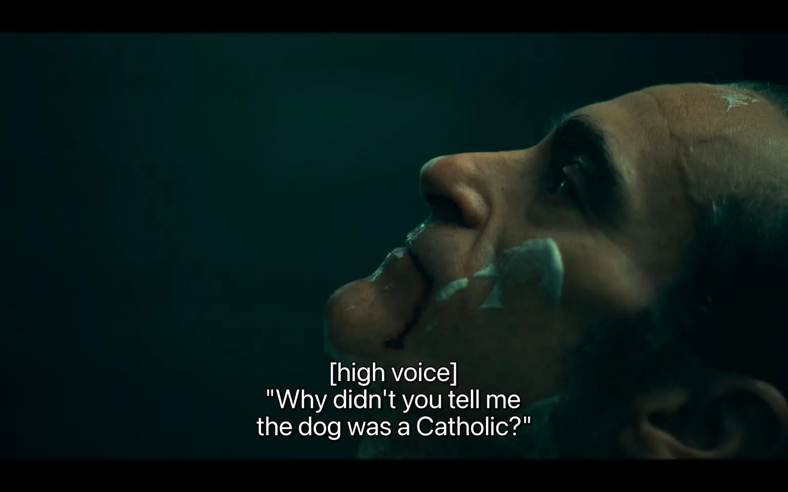 In the image, a character is depicted with a thoughtful expression, gazing upward. The lighting creates a moody atmosphere, with dark green tones dominating the background. The character has facial hair and appears to have some cream or substance applied to their face, possibly indicating a moment of vulnerability or reflection. The visible text in the image features a humorous quote, "Why didn't you tell me the dog was a Catholic?" spoken in a high voice, suggesting a comedic or absurd context. The overall scene conveys a blend of seriousness and humor, likely from a dramatic or comedic narrative.