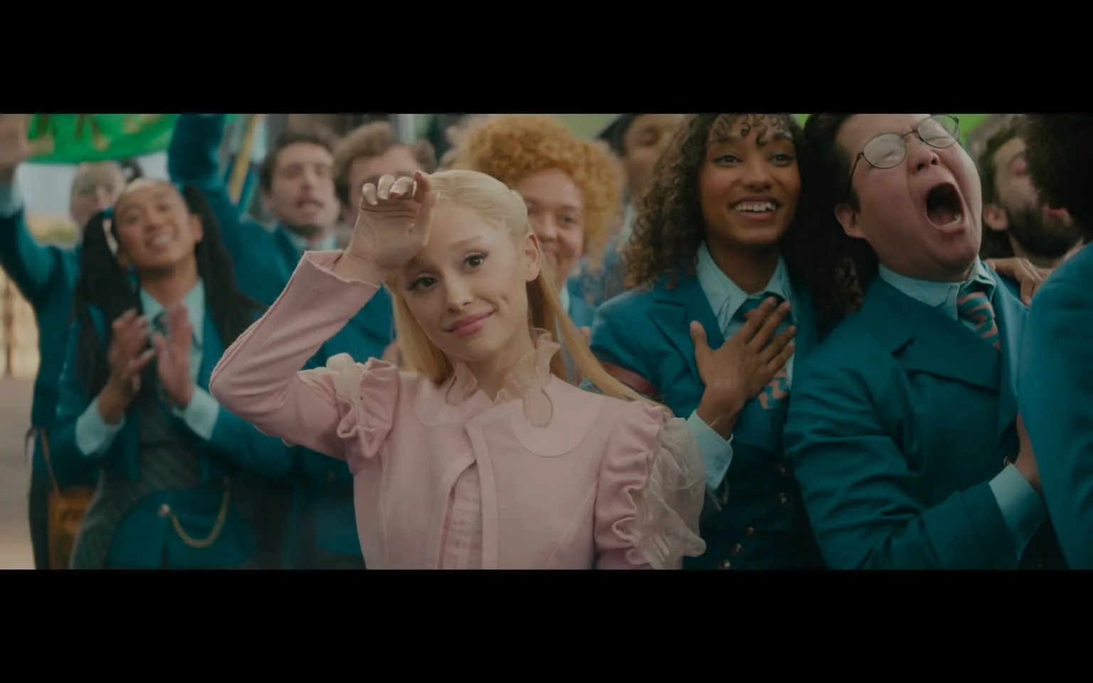 In a lively outdoor setting, a crowd is gathered, celebrating an event. The main subject, dressed in a light pink outfit with ruffled details, stands out among the group. She has long, straight blonde hair and is smiling, appearing to wave or greet the crowd. Surrounding her are several individuals, many wearing matching teal outfits, expressing joy and excitement. Some are clapping, while others have their hands over their hearts, indicating a sense of admiration or appreciation. The background features a green banner, adding to the festive atmosphere. The overall mood is cheerful and supportive, with a diverse group of people present, showcasing a range of hairstyles and expressions.