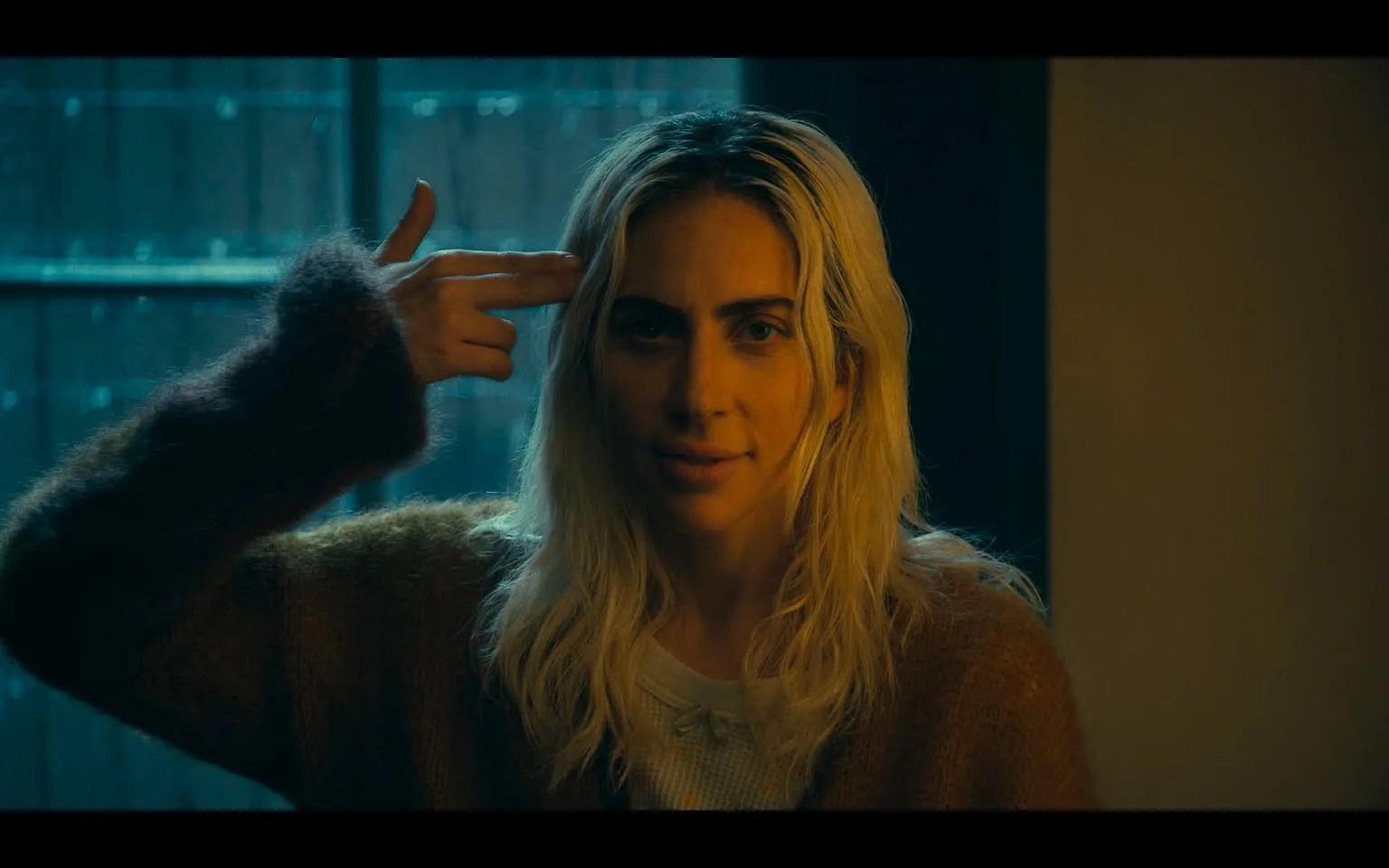 In the image, an unidentified adult with long, wavy blonde hair is positioned in a dimly lit indoor setting. They are wearing a light-colored top beneath a fuzzy, oversized brown sweater. The individual is looking directly at the camera with a slight smile, while their right hand is raised to their temple in a gesture that mimics a gun. The background features a window with a dark, possibly rainy exterior, contributing to a somber atmosphere. The lighting casts a blueish hue, enhancing the emotional weight of the scene. The gesture and expression may evoke themes of distress or irony, suggesting a complex emotional state.
