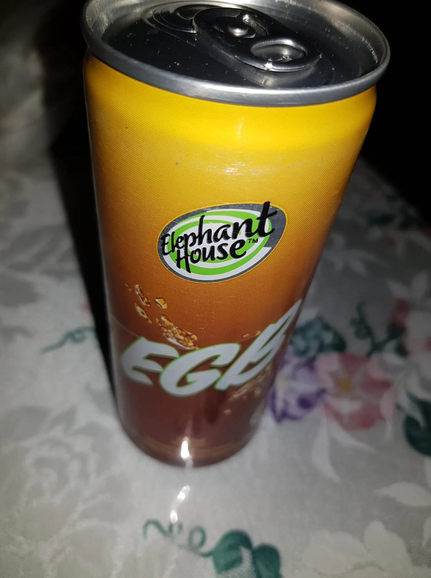 A beverage can is prominently displayed in the image, featuring a gradient design that transitions from yellow at the top to a darker shade at the bottom. The can is labeled with the brand name "Elephant House," which is visible in a circular logo. The text "EGE" is also present, likely indicating the type of beverage. The background is a patterned tablecloth with floral designs, suggesting an indoor setting. The lighting appears dim, adding a subtle ambiance to the scene. There are no visible people or any sensitive content in the image.