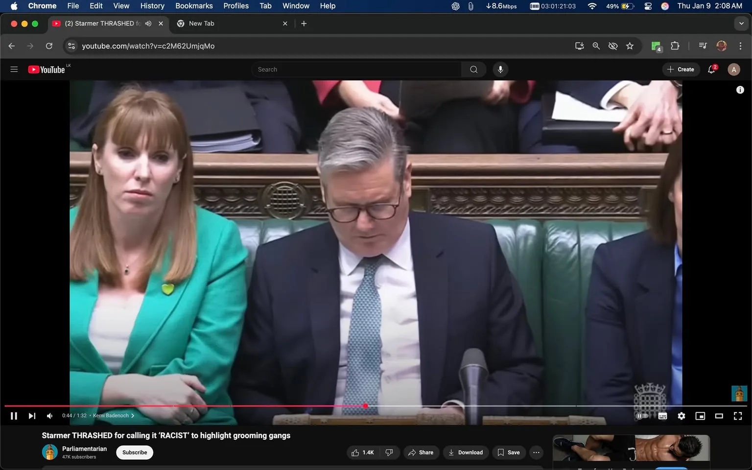 In a parliamentary setting, two individuals are seated on a green leather bench. The person on the left has long, straight hair and is wearing a bright green blazer with a yellow pin. They appear to be listening intently, with a serious expression. The central figure, dressed in a dark suit and glasses, is looking down, seemingly focused on something in their hands. The background shows other individuals, some holding papers, indicating a formal discussion or debate is taking place. The atmosphere appears tense, reflecting the serious nature of parliamentary proceedings.