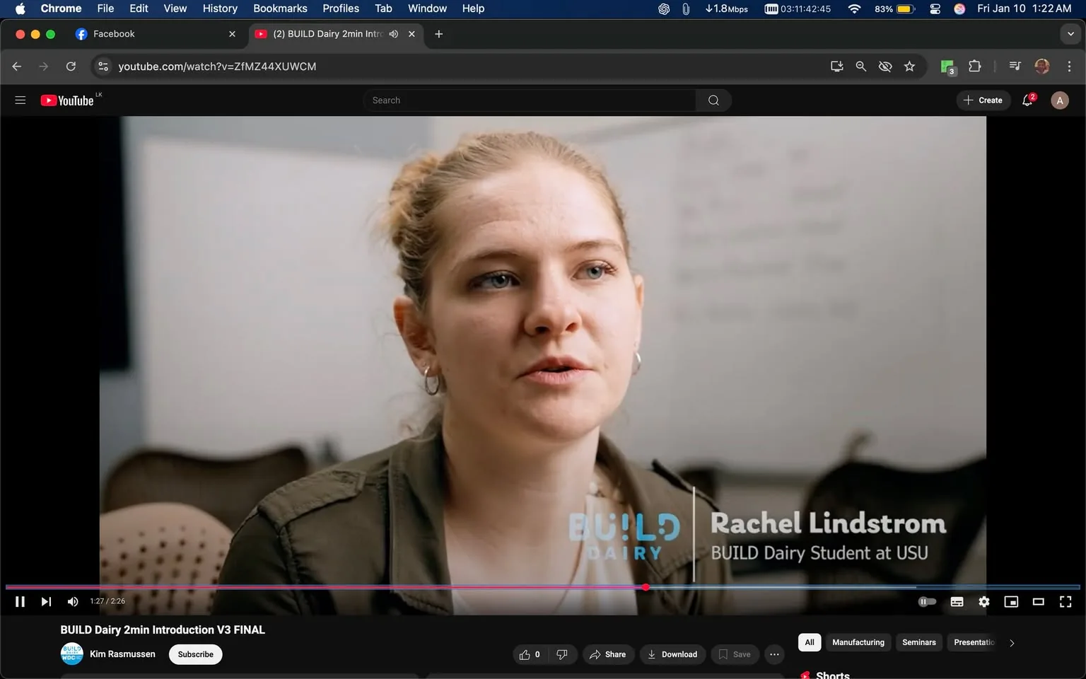 In the image, a young adult woman is speaking directly to the camera in a professional environment. She has light brown hair styled in a loose bun and is wearing a dark green jacket over a white top. Her expression appears focused and engaged as she discusses her experiences. The background is slightly blurred, but there are hints of a classroom or meeting space, with some chairs visible. Prominently displayed on the screen is the logo "BUILD Dairy," along with her name, Rachel Lindstrom, and her title as a BUILD Dairy student at USU. The overall atmosphere suggests an informative or promotional video.