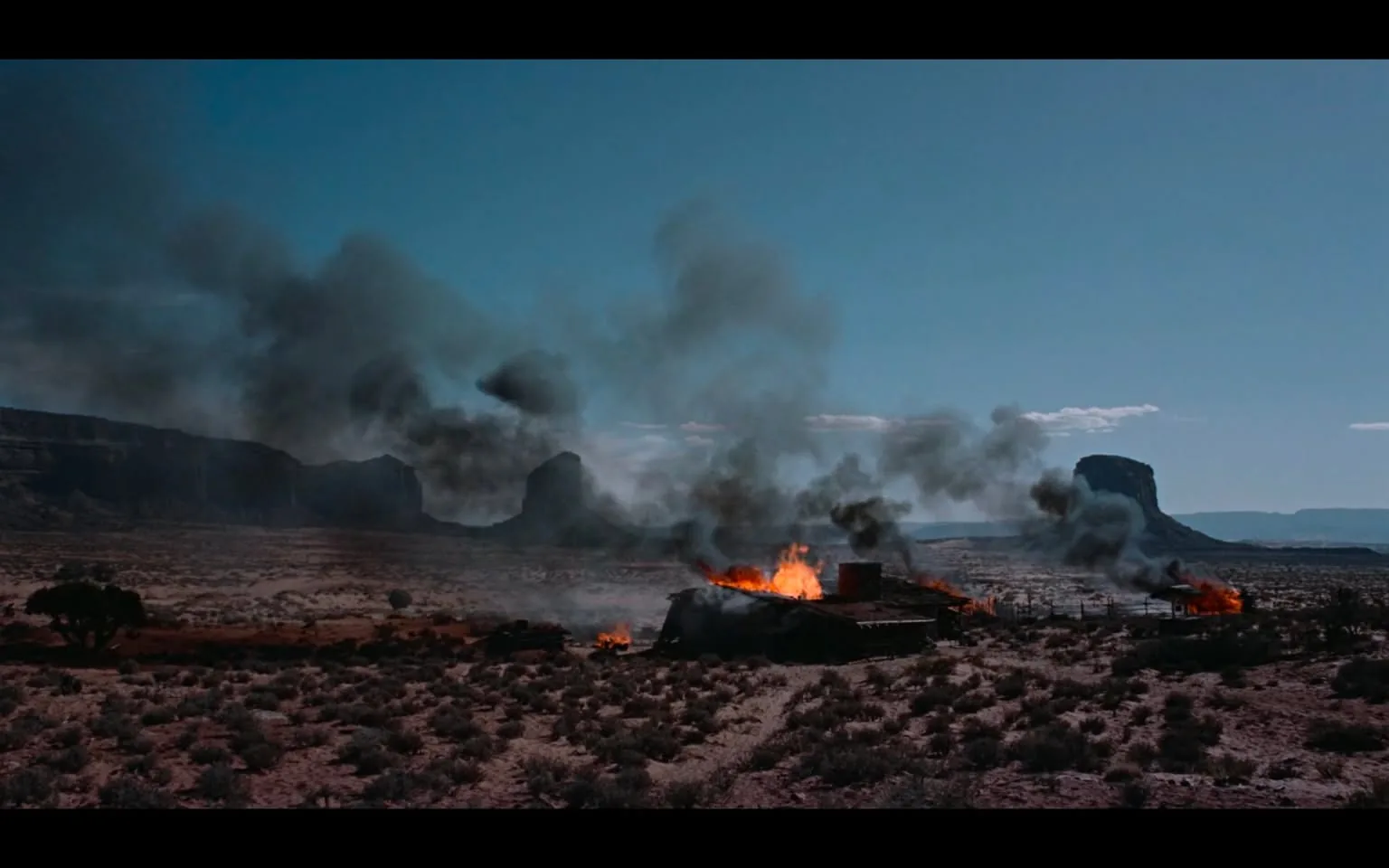 In a stark, arid landscape, a structure is engulfed in flames, sending thick plumes of black smoke into the clear blue sky. The fire appears to be consuming the building, which is situated amidst sparse vegetation and rocky formations in the background. The terrain is dry and rugged, with distant mountains adding to the dramatic scenery. The scene evokes a sense of chaos and destruction, suggesting a violent event has occurred. There are no visible people or identifiable brands in the image, focusing solely on the aftermath of the fire.