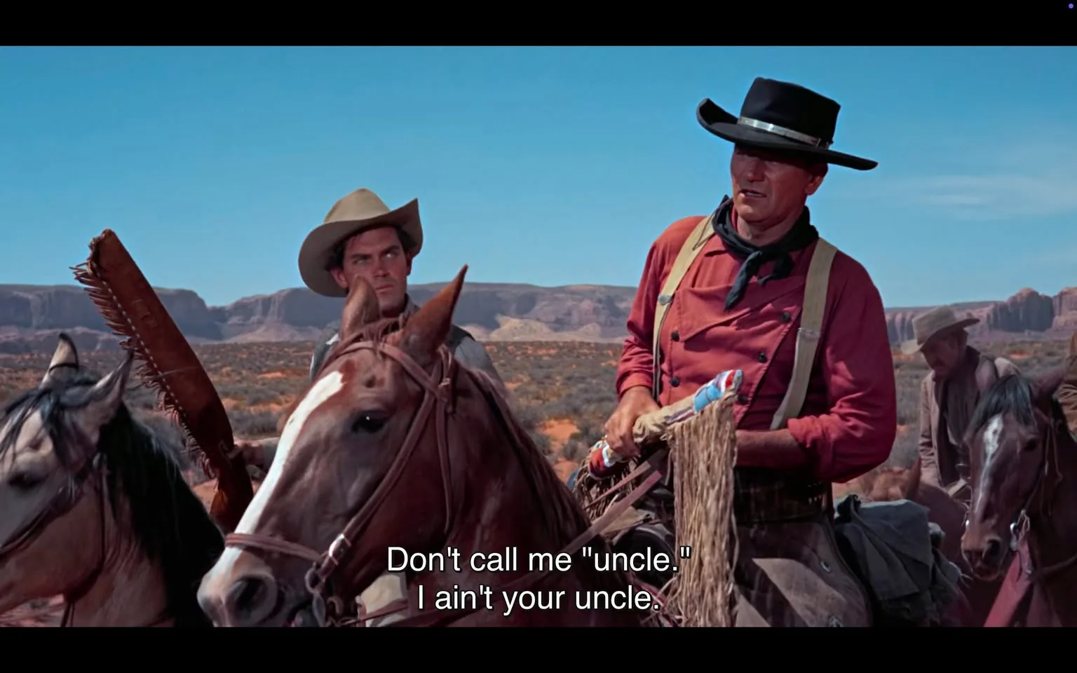 In the image, two men are mounted on horses in a vast desert landscape, characterized by red rock formations and a clear blue sky. The man on the right, dressed in a red shirt and a black hat, appears to be delivering a line of dialogue, stating, "Don't call me 'uncle.' I ain't your uncle." The other man, wearing a light-colored cowboy hat, looks on attentively. Both men are equipped with saddles and appear to be engaged in a conversation, suggesting a narrative moment. The setting is outdoors, likely during the day, with no visible signs of violence or sexual content.