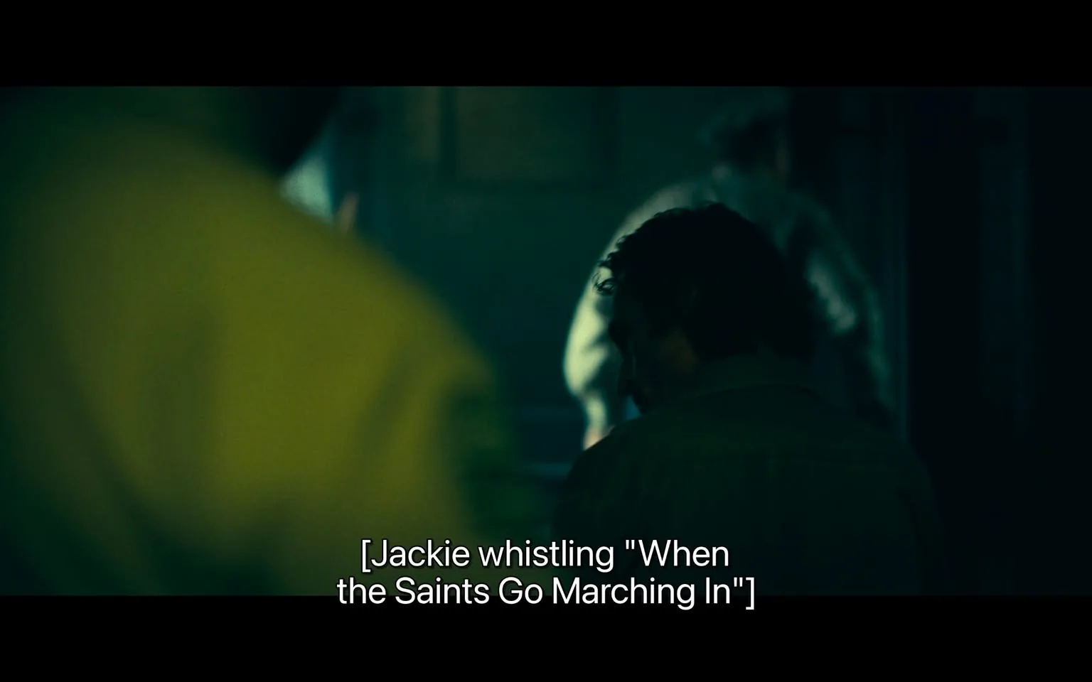 In the scene, a character is seen from behind, with dark hair and wearing a light-colored shirt. The atmosphere is dimly lit, creating a moody ambiance. The character appears to be whistling the tune "When the Saints Go Marching In," suggesting a moment of reflection or lightheartedness amidst a possibly tense setting. In the background, there are blurred figures, indicating that the scene is likely set indoors, possibly in a bar or a similar environment. The overall tone is somber, with shadows and muted colors dominating the visual.