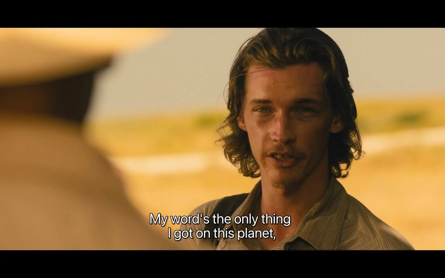 In the scene, an unidentified young adult male is speaking earnestly, expressing the sentiment that his word is his most valuable asset. He has long, wavy hair and a light mustache, wearing a button-up shirt with a subtle pattern. The background is an outdoor setting, likely a rural area, with a blurred view of a field or landscape behind him, suggesting a warm, sunny day. The dialogue displayed on the screen reads, "My word's the only thing I got on this planet," indicating a moment of personal significance or a serious conversation. The focus is on the character's expression, which conveys sincerity and determination.