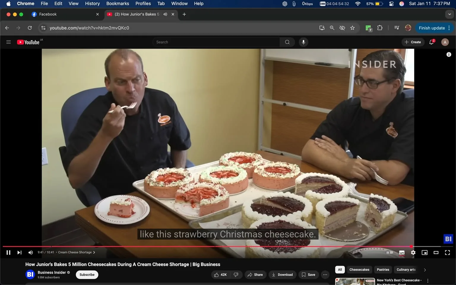 In the image, two adults are seated at a table in a casual indoor setting. One individual, wearing a black chef's jacket with the "Junior's" logo, is tasting a slice of cheesecake with a fork, displaying a thoughtful expression. The other adult, also in a black chef's jacket, watches attentively. On the table, there is a tray filled with various cheesecakes, including a strawberry cheesecake prominently featured, decorated with whipped cream and a red topping. The background includes a simple office-like environment, with a light-colored wall and a wooden table. The scene conveys a relaxed atmosphere focused on the enjoyment of desserts.