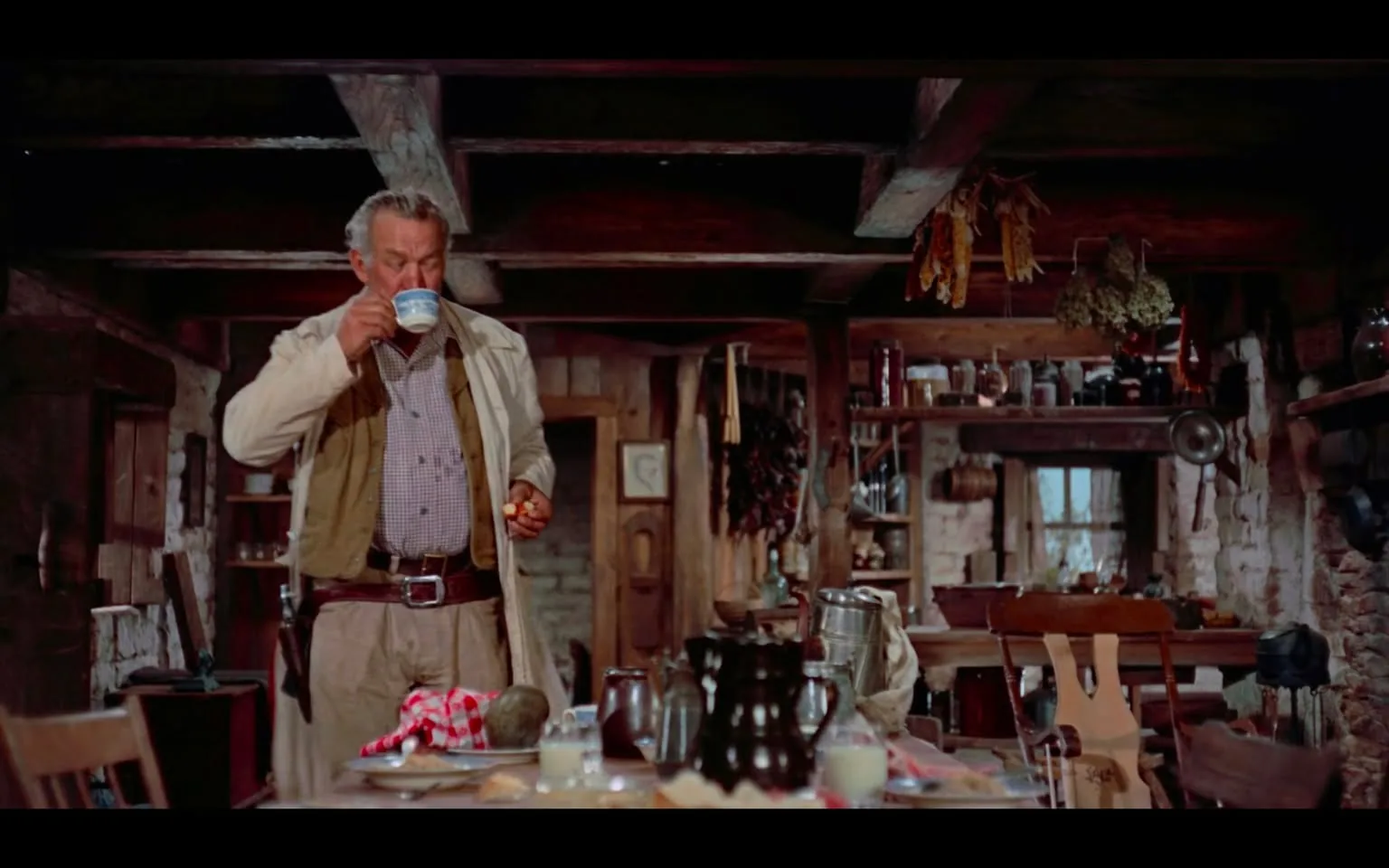 In a rustic kitchen, an unidentified adult man stands at a table, sipping from a blue cup. He wears a light-colored jacket over a checkered shirt and beige pants, with a belt that has a visible buckle. In his other hand, he holds a small piece of food. The kitchen is filled with wooden beams and shelves lined with jars and various kitchen items, creating a warm, homey atmosphere. On the table, there are several food items, including bread and a checkered cloth. The setting appears cozy and lived-in, suggesting a rural or traditional environment.