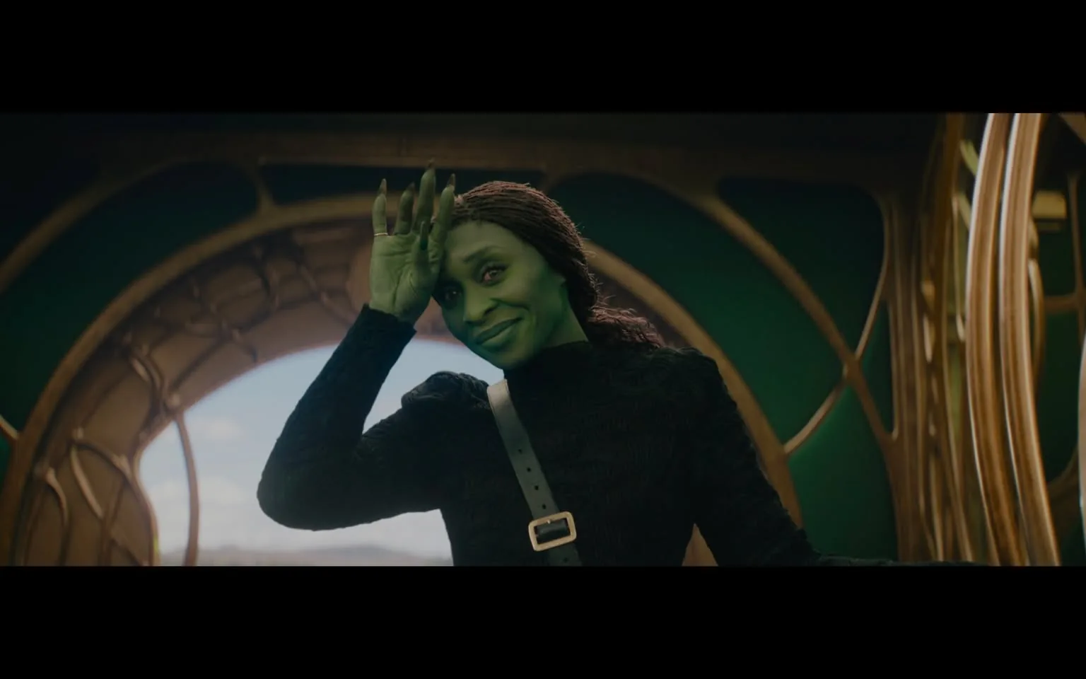 In the image, a character with green skin and a warm smile is seen waving. They have long, dark hair styled in loose waves and are wearing a black, textured outfit with a belt featuring a prominent buckle. The background is an indoor setting with a circular, ornate design, suggesting a fantastical or sci-fi environment. The character's expression is friendly and inviting, contributing to a lighthearted atmosphere. The overall composition hints at a scene from a fictional narrative, likely involving themes of adventure or camaraderie.